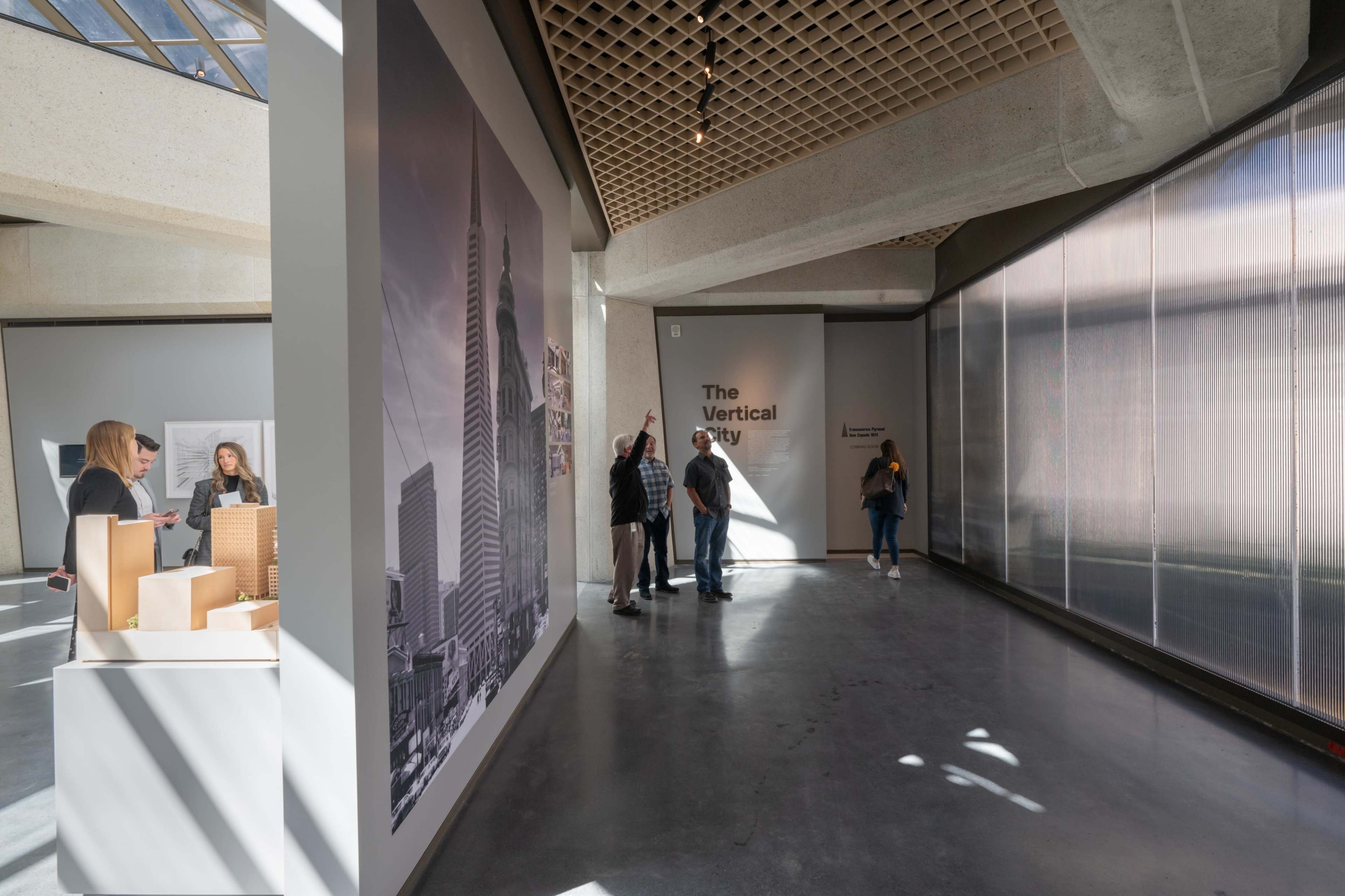 The image shows a spacious exhibition area featuring a large wall graphic of a skyscraper, a sign reading "The Vertical City," and several visitors engaging with the displays.
