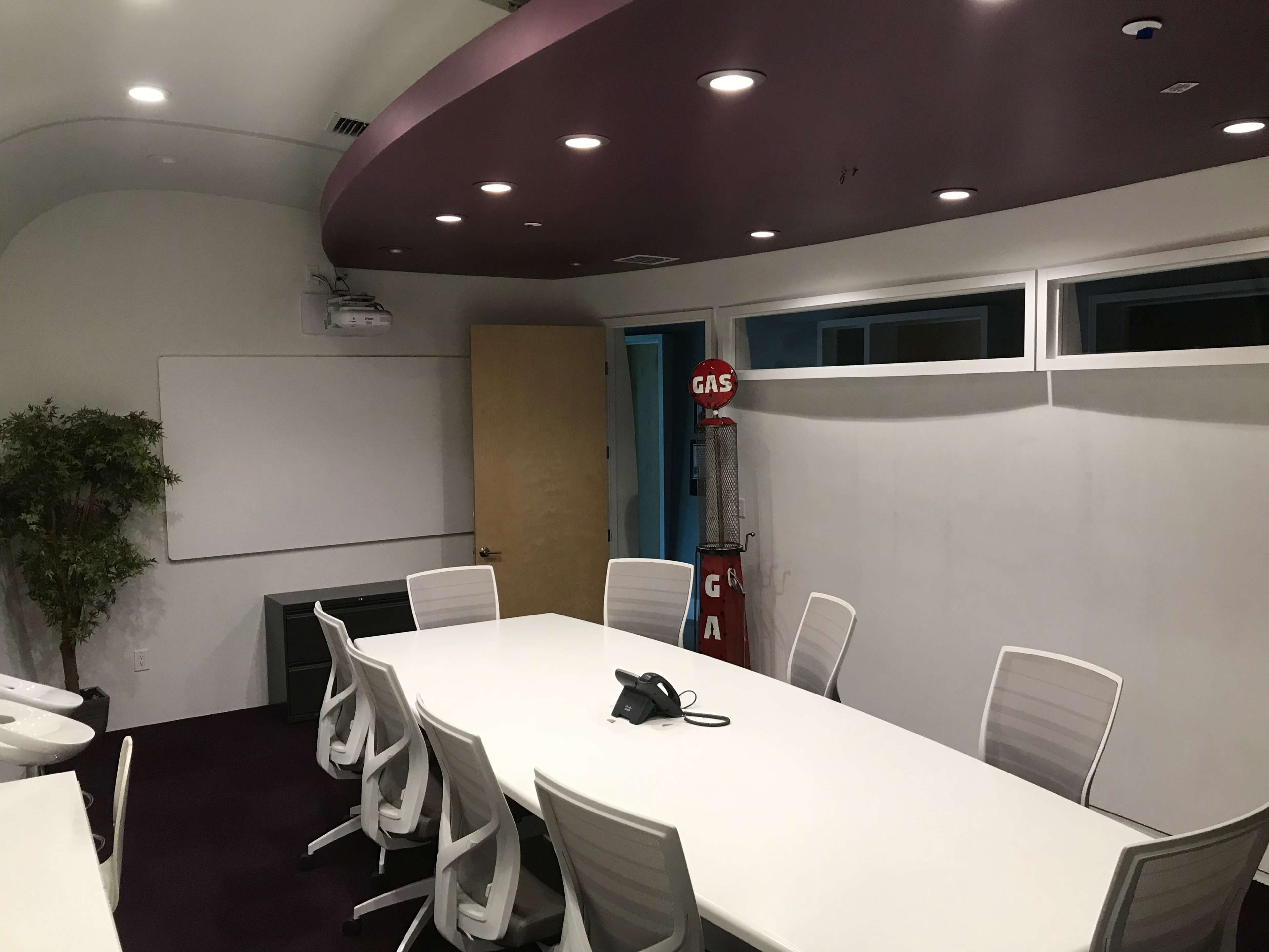 A conference room features a long white table surrounded by several white chairs, a projector mounted on the ceiling, and a whiteboard on a wall.