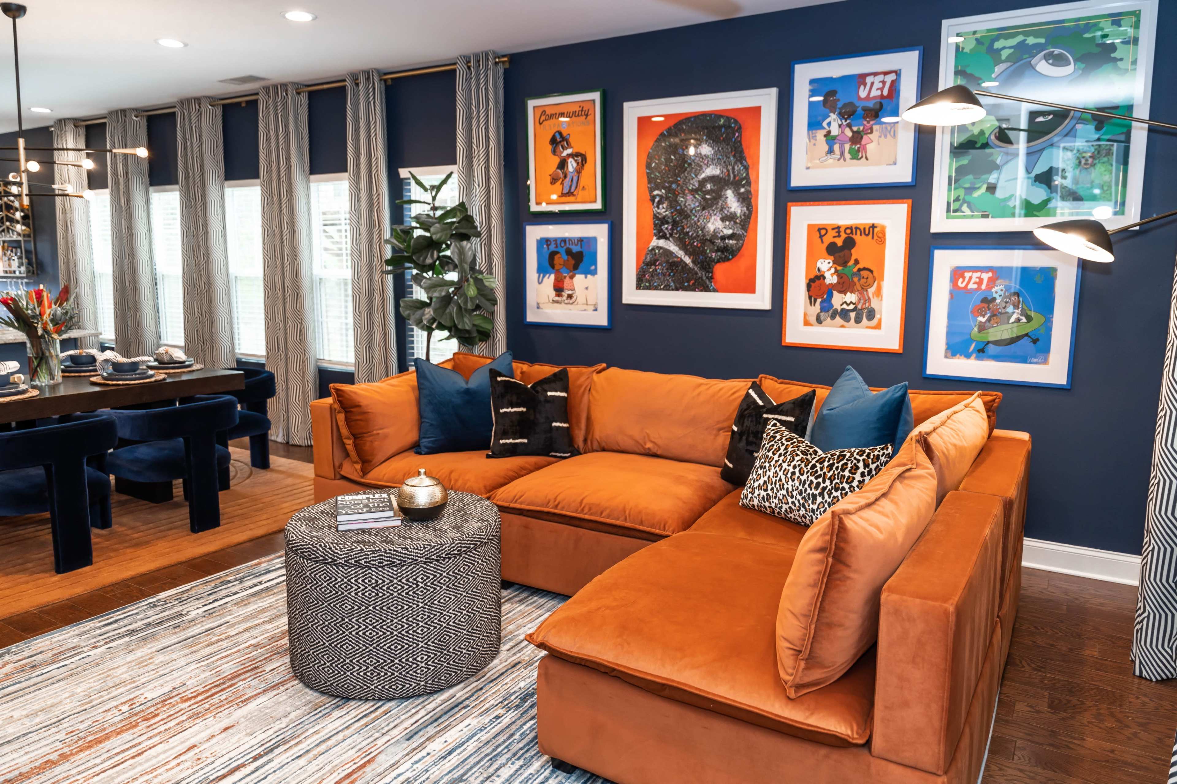A modern living room features a large orange sectional sofa, a round patterned coffee table, and framed colorful artwork on the blue walls.