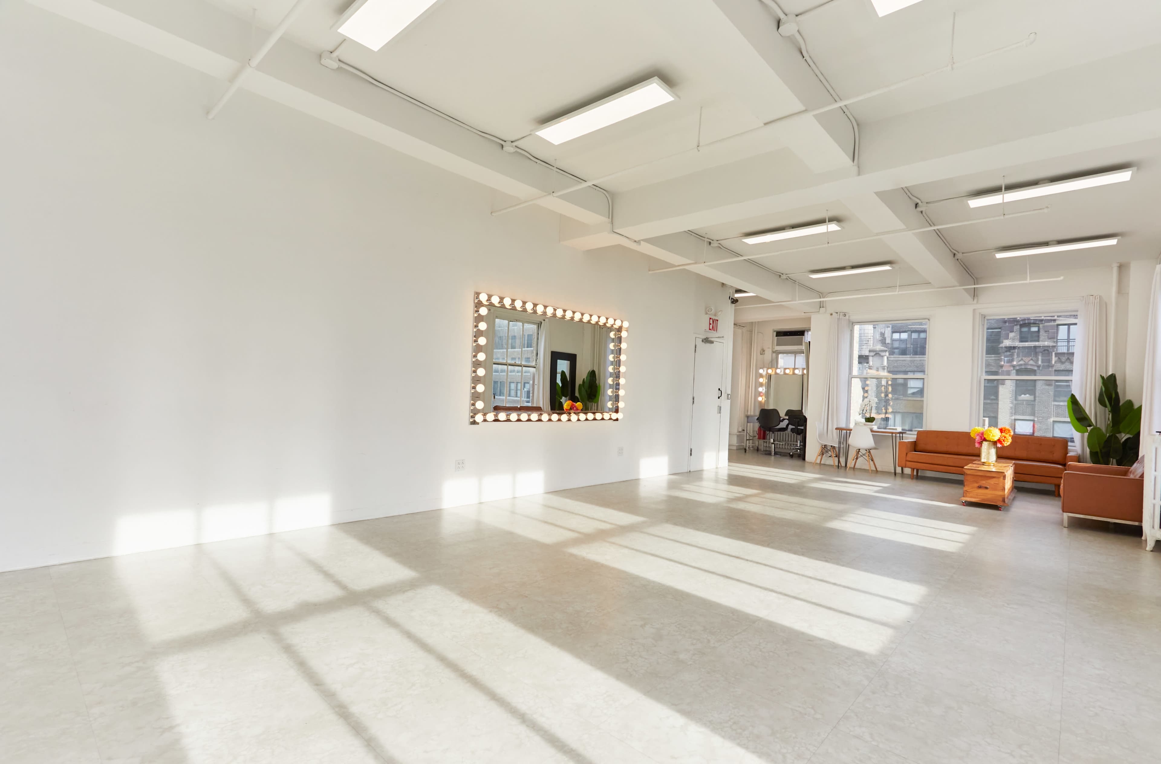 The image shows a spacious, well-lit room with white walls, a large mirror surrounded by light bulbs, and two seating areas, including a brown couch and a wooden table with flowers.