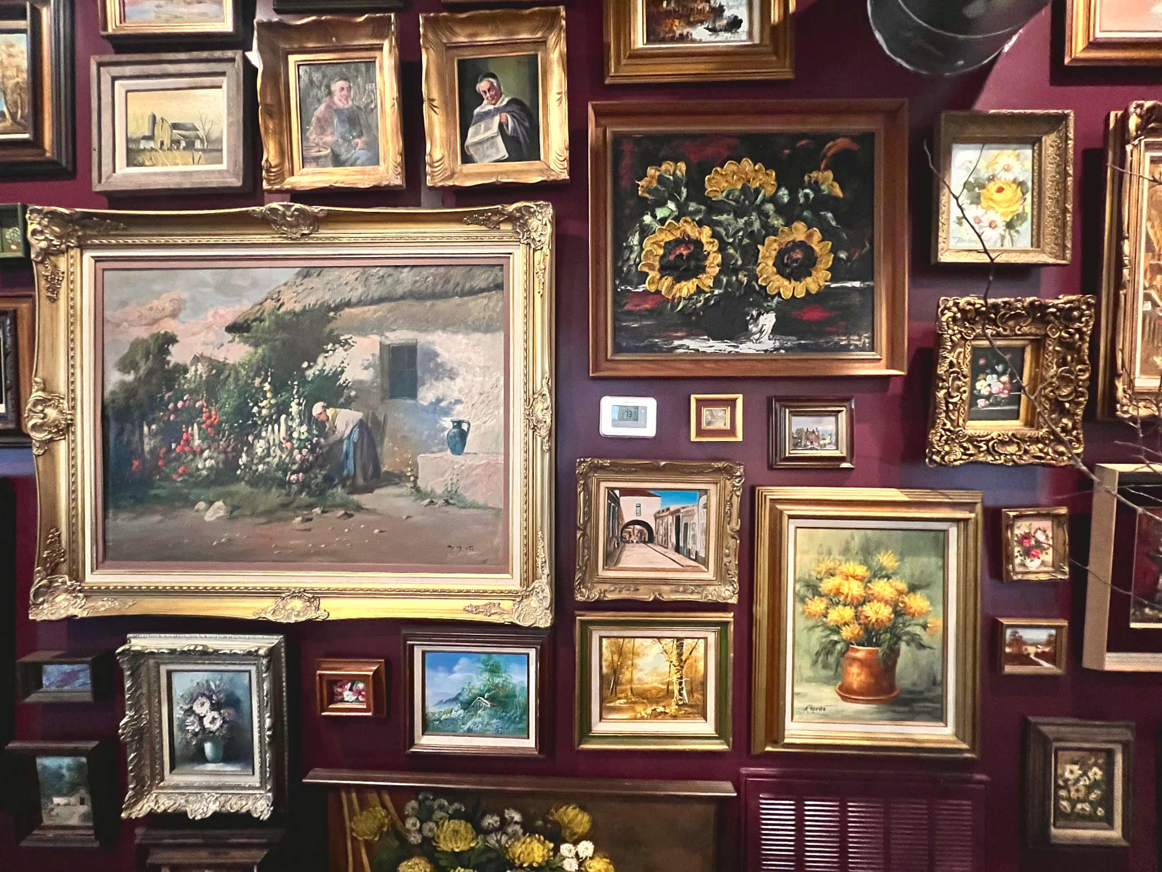The image shows a collection of framed paintings displayed in a gallery setting with a burgundy wall as the backdrop.