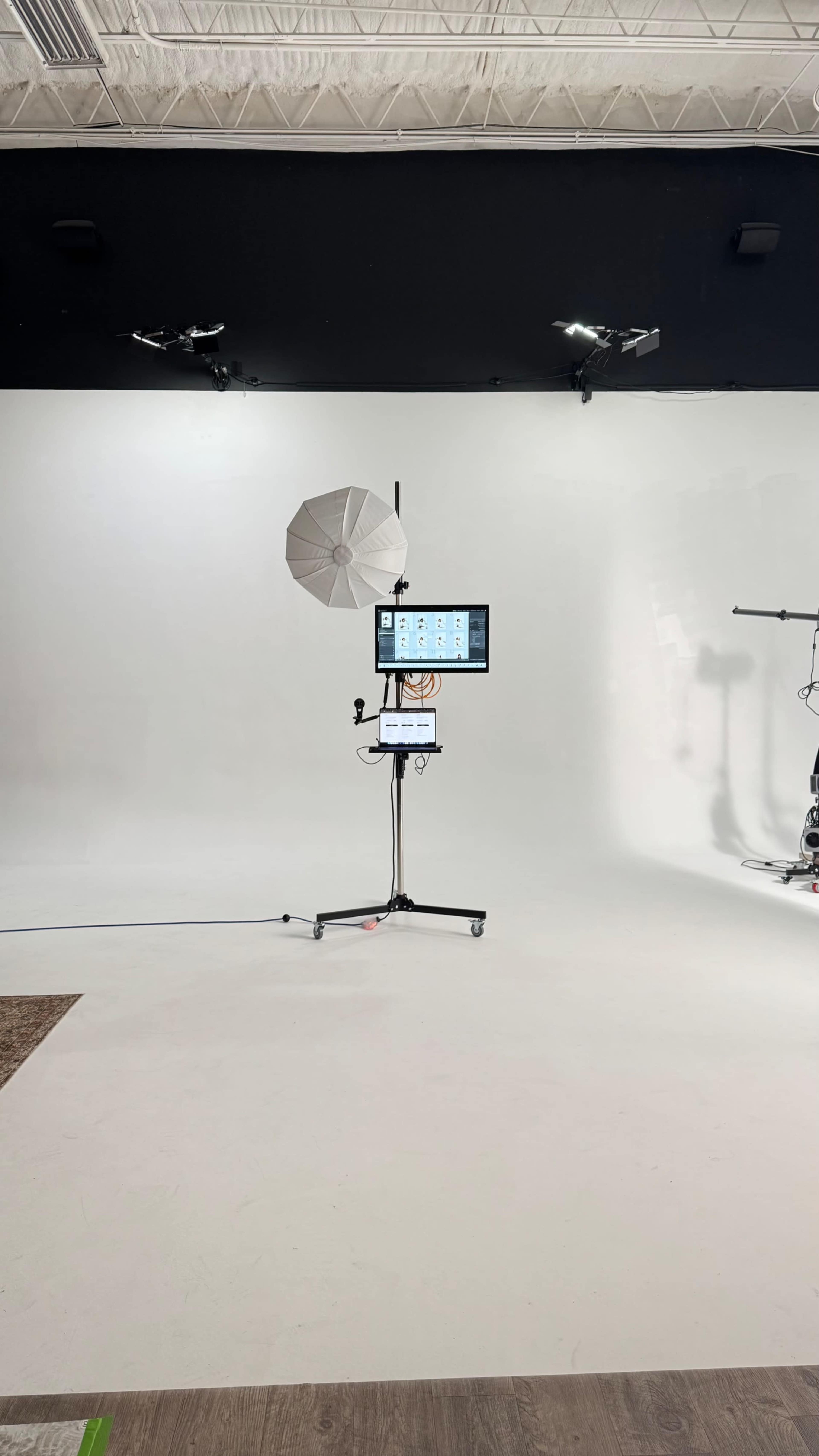 The image shows a photo studio setup featuring a monitor on a stand, a softbox light, and various studio equipment in a space with white walls and a smooth floor.