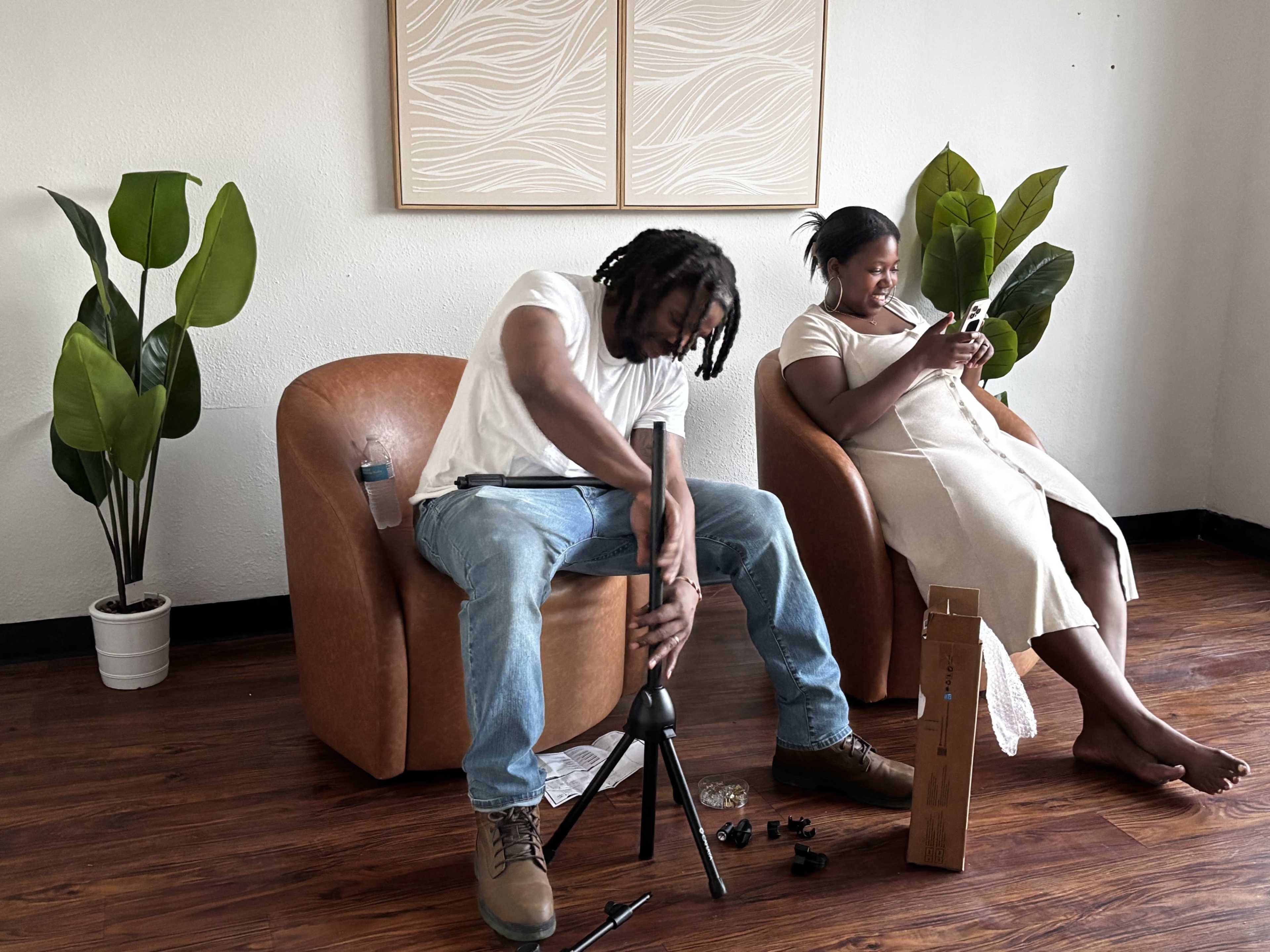 A young man adjusts a camera tripod while a woman sits nearby, engrossed in her phone, in a room decorated with potted plants and minimalist artwork.