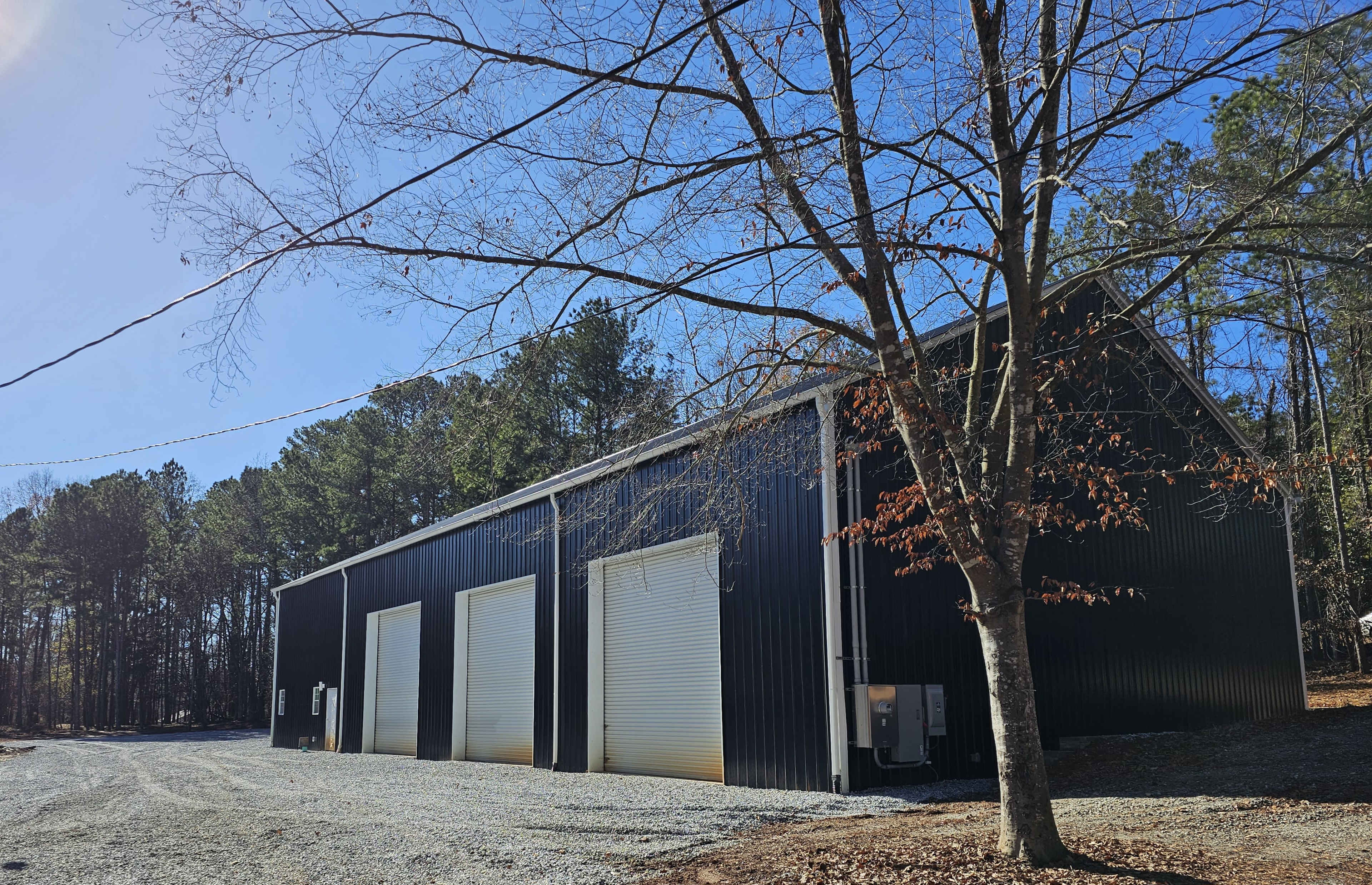 The image shows a black metal building with three large garage doors, surrounded by trees, on a gravel driveway under a clear blue sky.