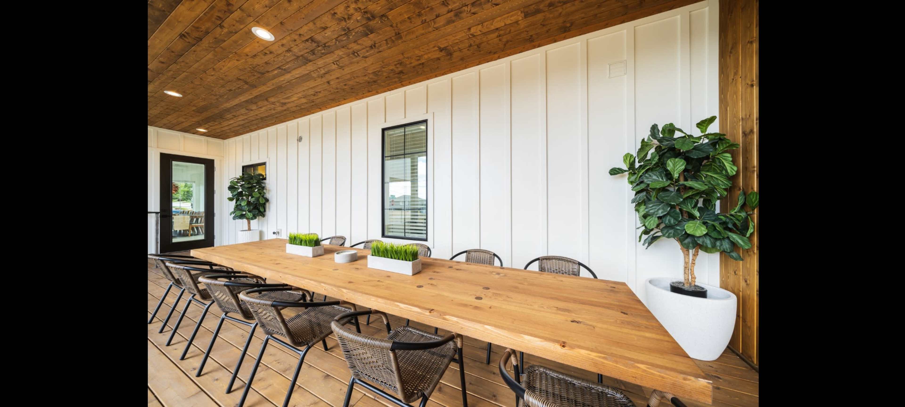 A spacious outdoor dining area featuring a long wooden table surrounded by wicker chairs, with potted plants on either side and a wooden ceiling overhead.