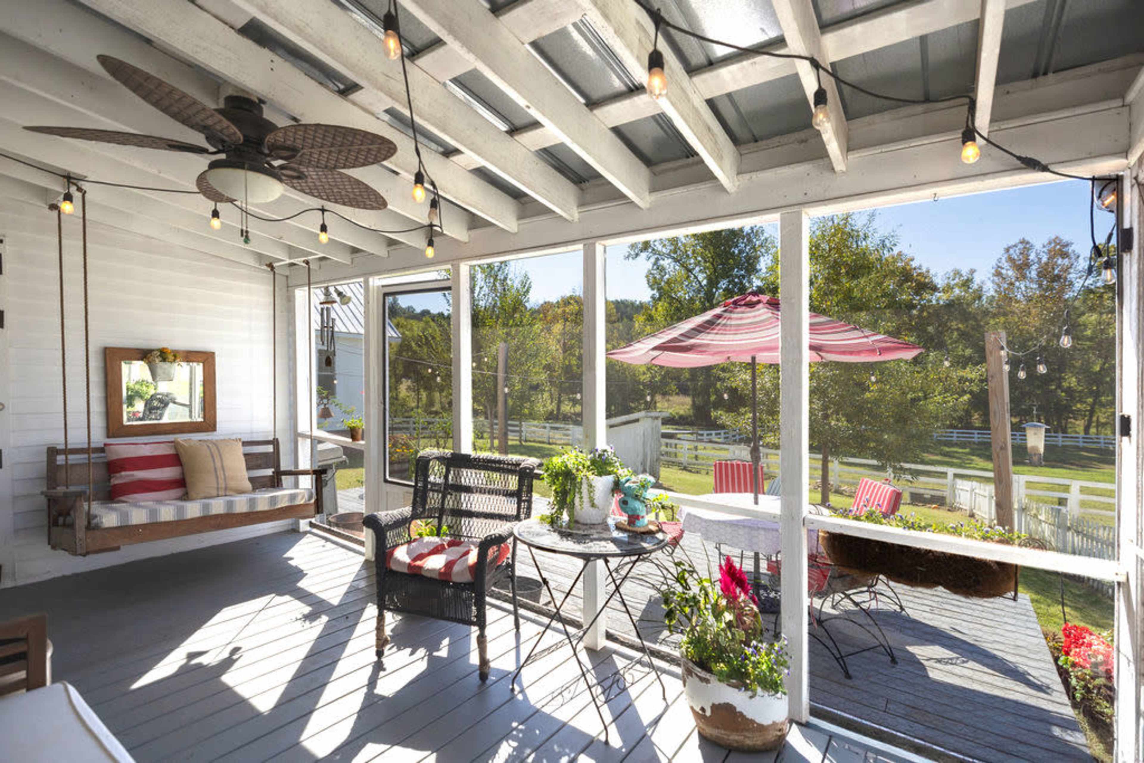 The image shows a screened-in porch with a ceiling fan, a swing bench, and a round table surrounded by chairs, overlooking a green outdoor landscape.