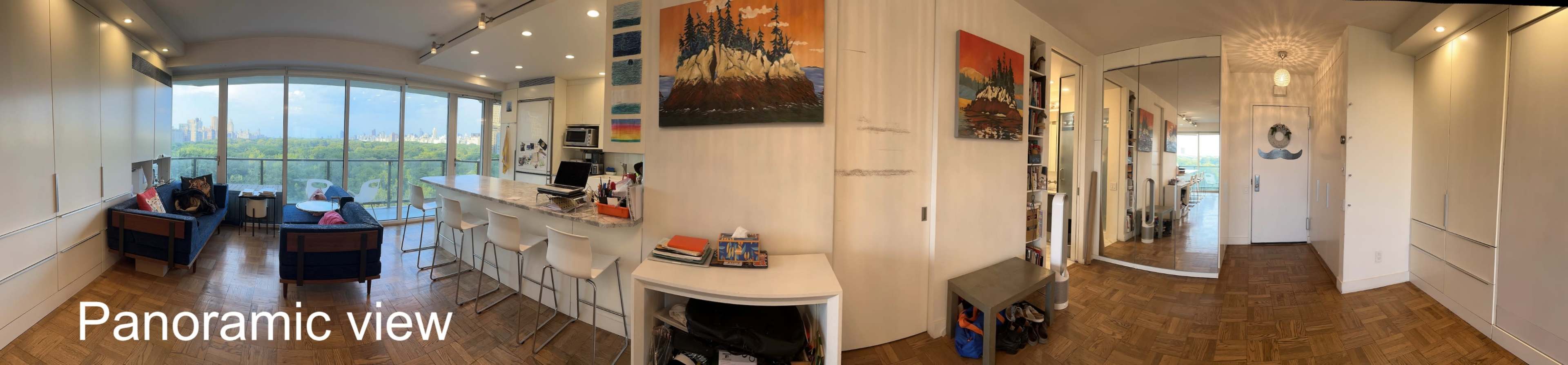 The image shows a panoramic view of a modern indoor space featuring a kitchen, living area with seating, and an entrance to a hallway.