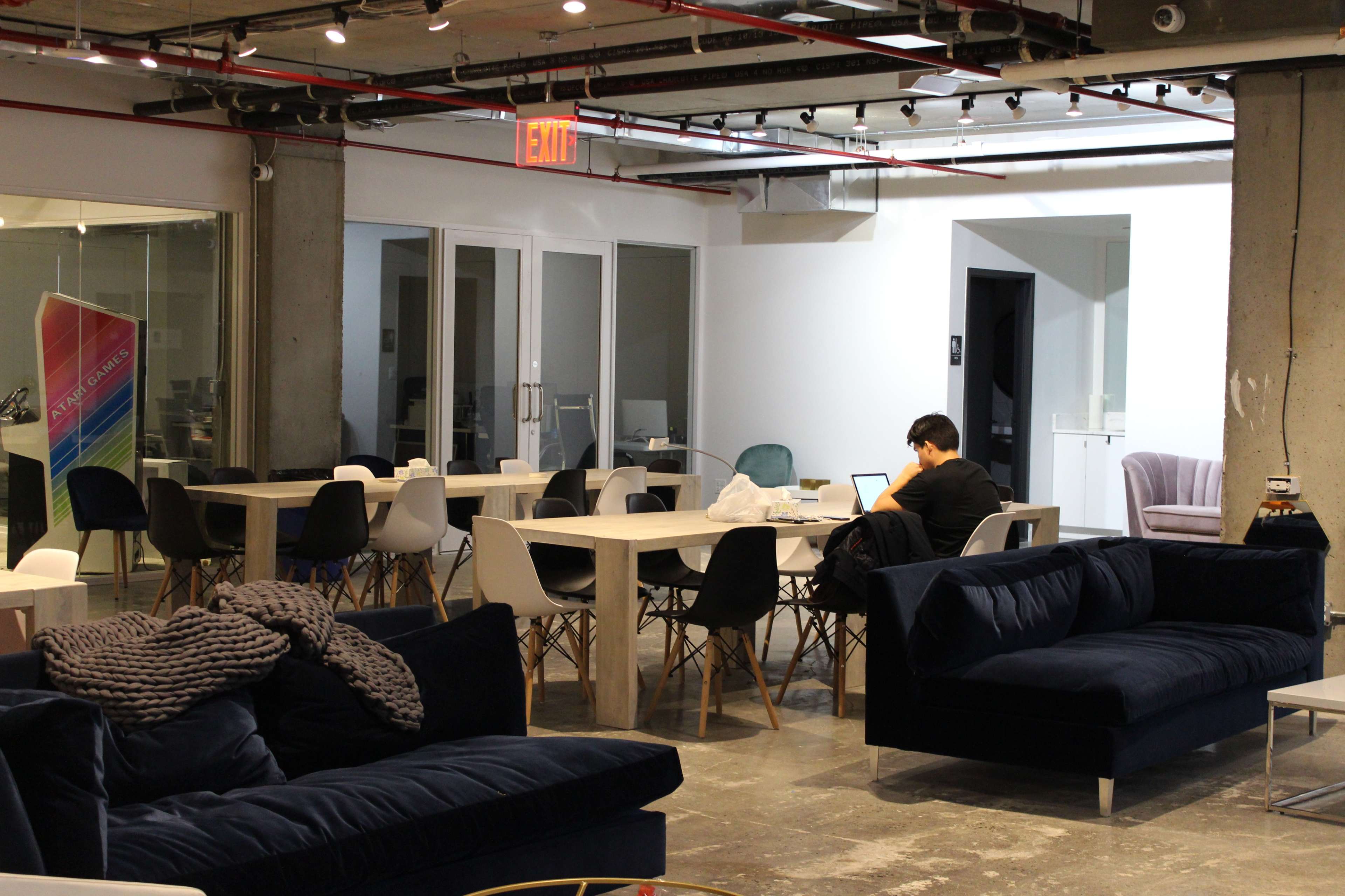 A person works on a laptop at a table in a modern open-office space with various seating areas and bright lighting.