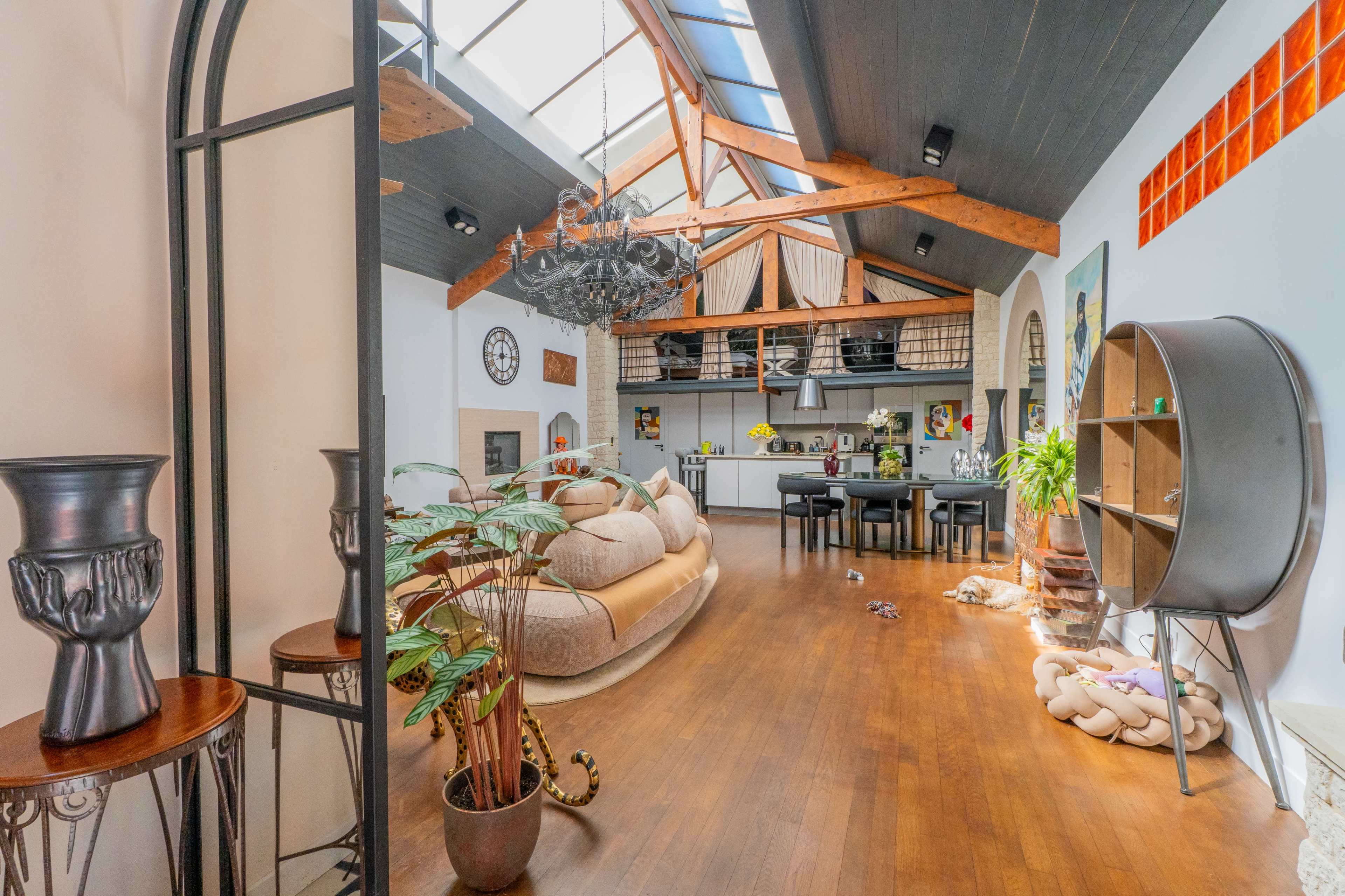 The image features a spacious, modern living area with high ceilings, wooden beams, and a large sofa, complemented by various plants and decorative elements.