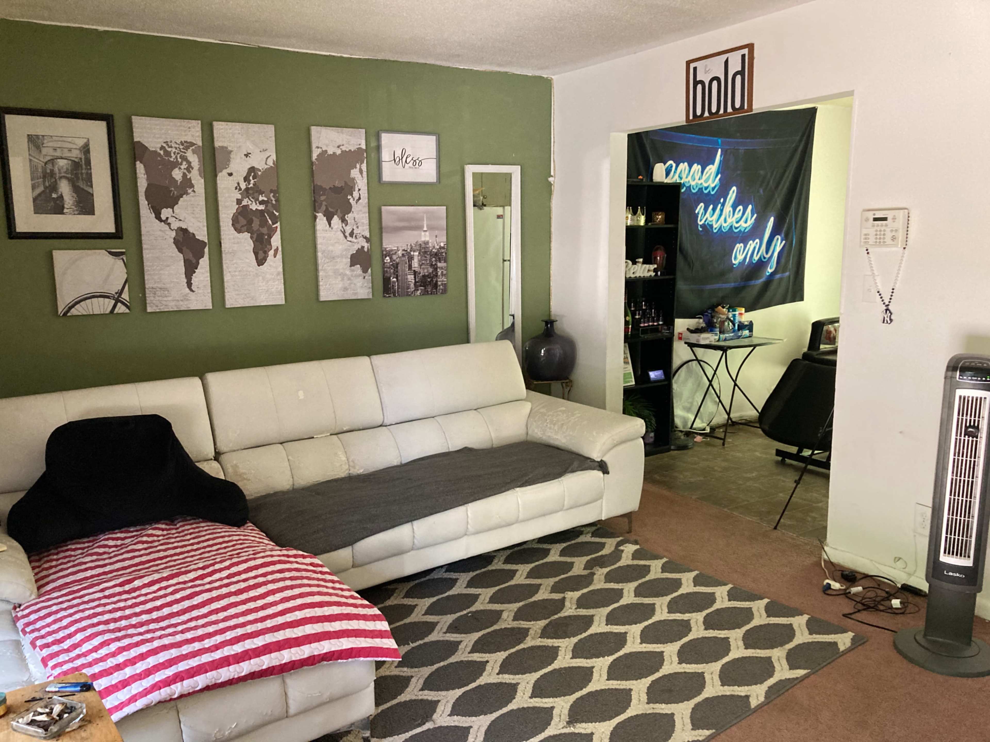 A living room with a white sofa, a striped throw blanket, a green accent wall, and framed world maps, leading to a room with a sign that reads "good vibes only."