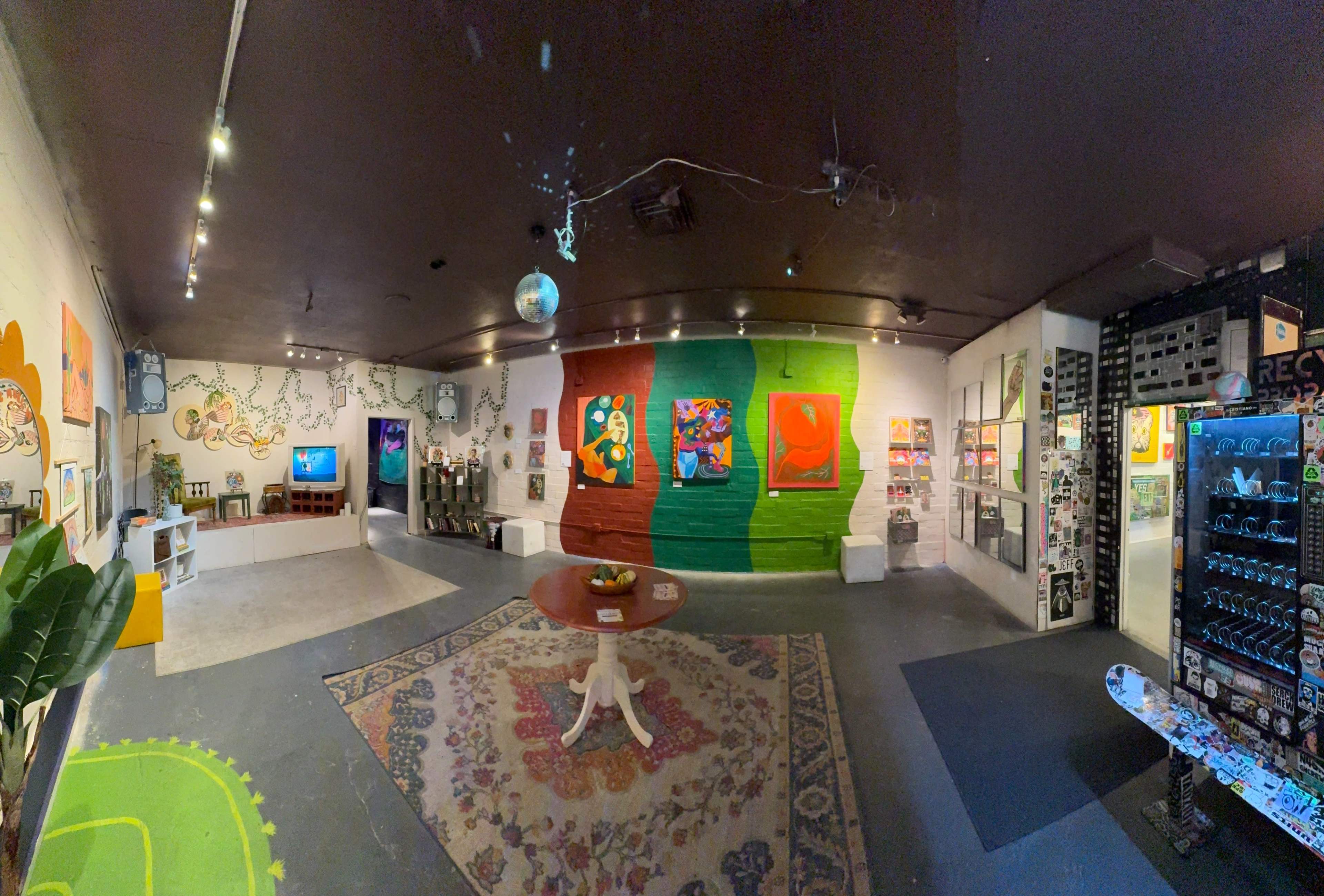 The image shows an art gallery interior featuring colorful paintings on the walls, a central table, and various decorative items throughout the space.