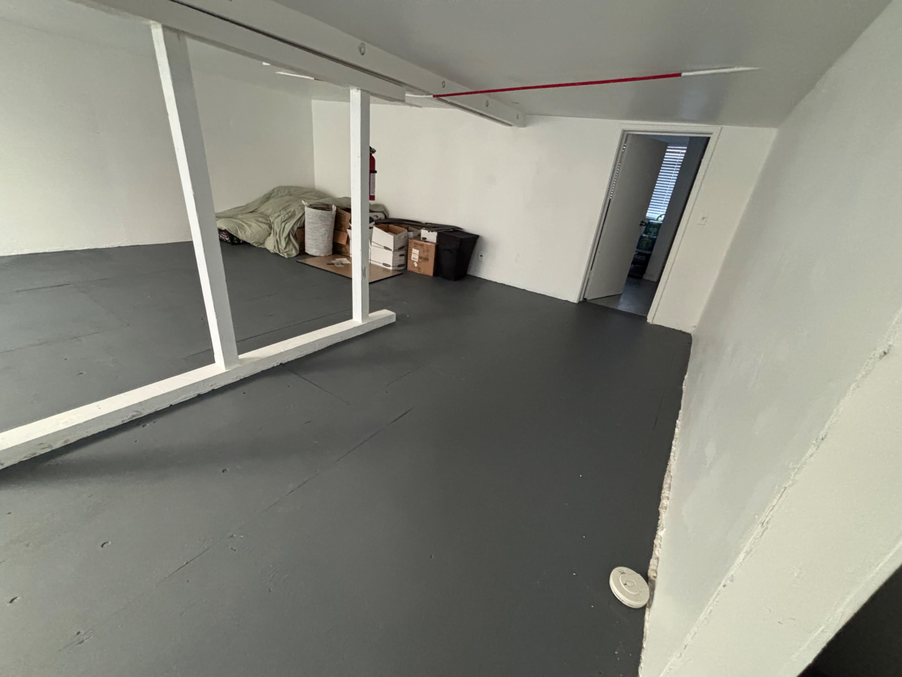The image shows an empty room with gray flooring, white walls, a partially visible door, and some boxes stacked along the wall.