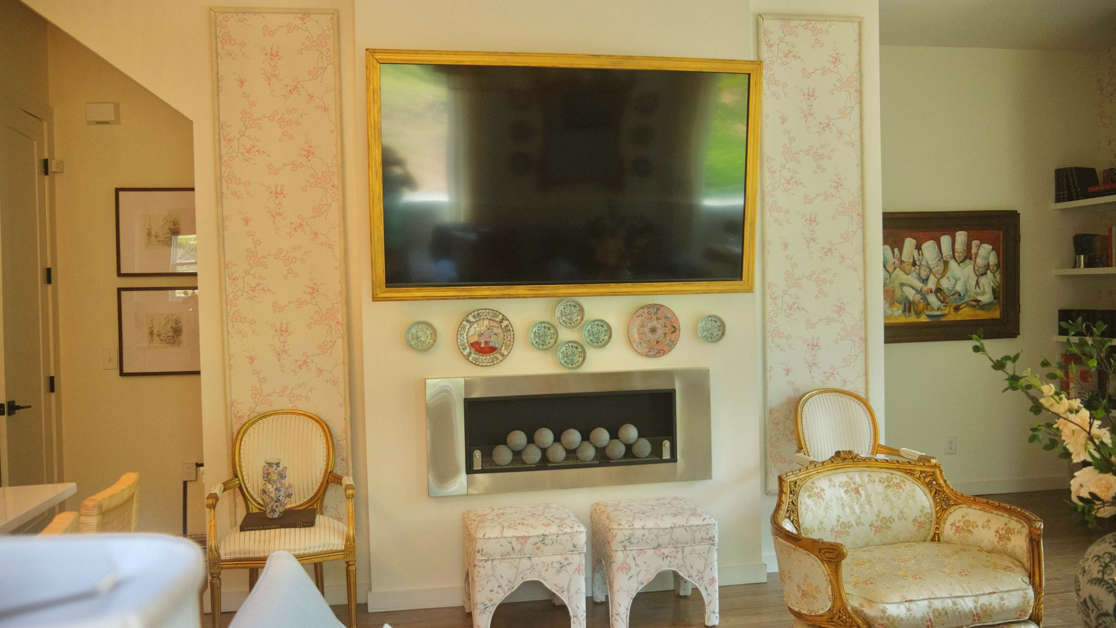 The image shows a living room with a large flat-screen TV mounted above a decorative fireplace, flanked by floral-patterned panels and accompanied by two ornate chairs and patterned stools.