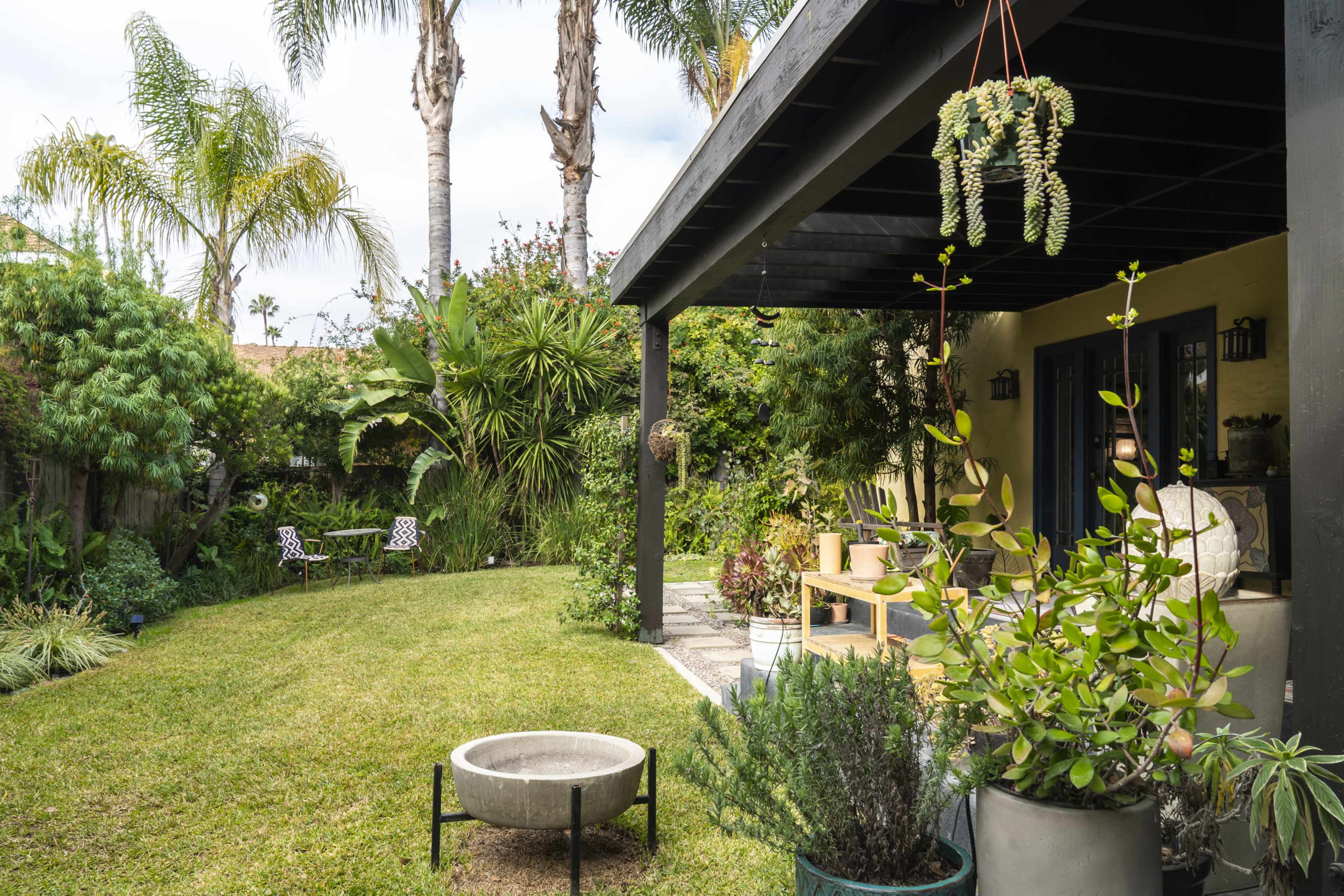 The image shows a well-maintained backyard with a lawn, various potted plants, and patio furniture under a covered area.