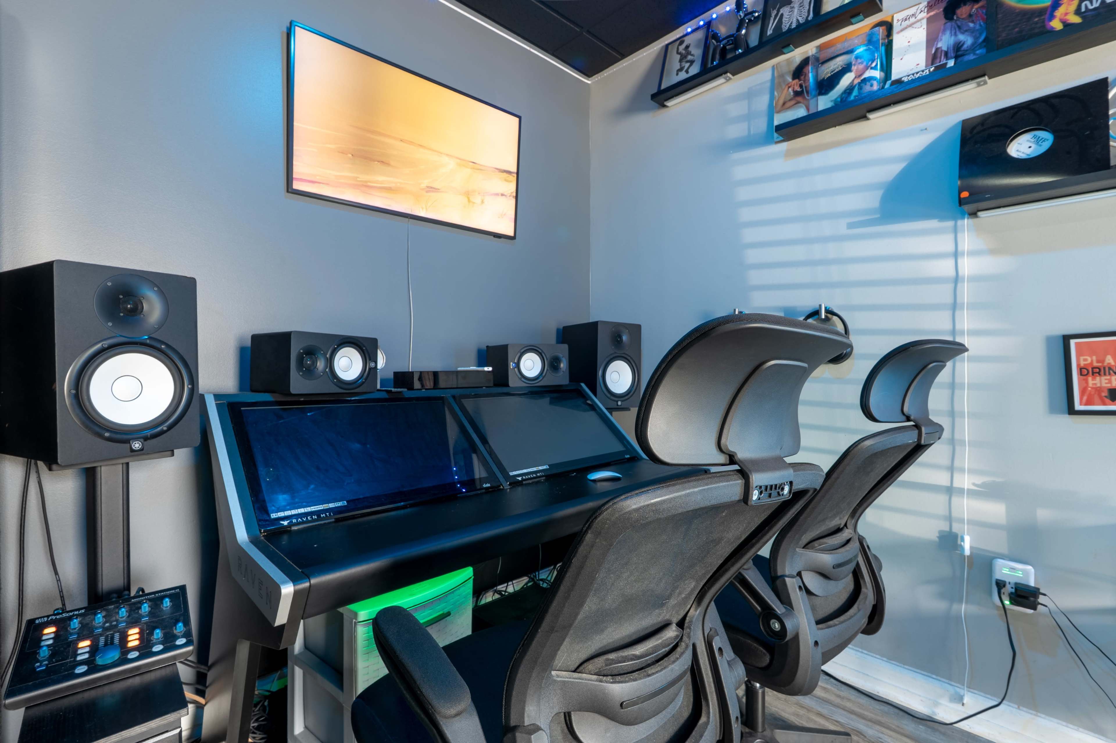 The image shows a modern audio production studio featuring two computer monitors, two office chairs, and audio speakers positioned on either side against a gray wall.
