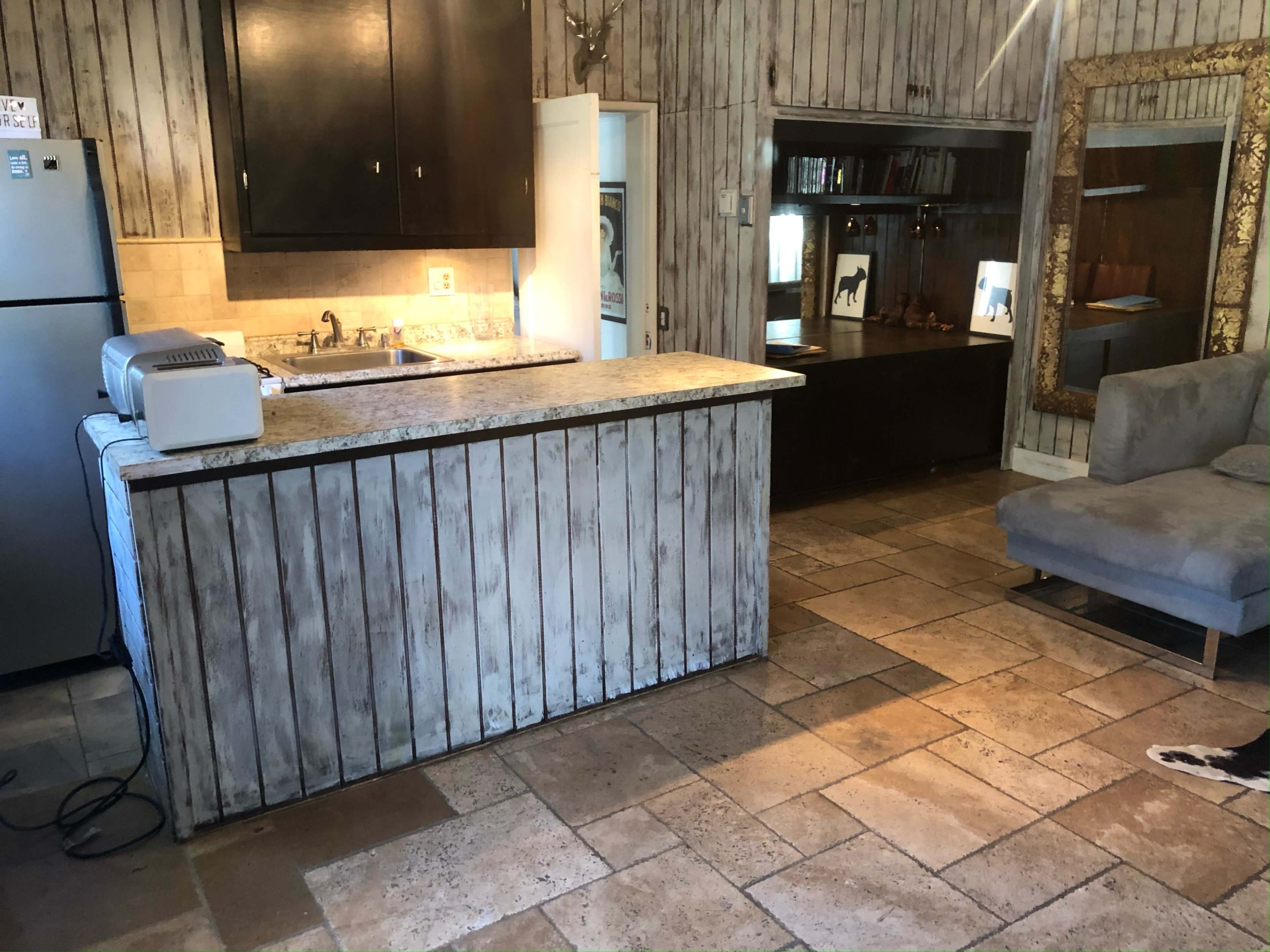 The image shows a rustic kitchen area with a stone-tiled floor, a wooden counter, and a cozy seating section featuring a gray couch.