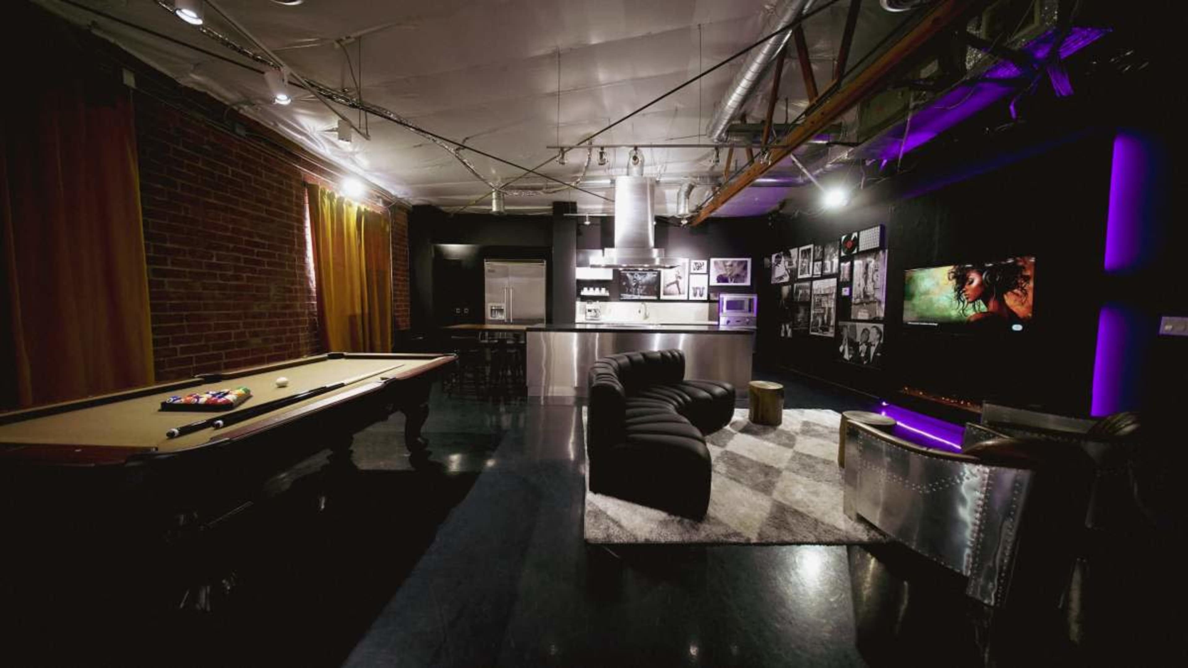 The image shows a modern lounge area featuring a pool table, a sleek kitchen, and a comfortable seating arrangement alongside a wall of photographs.