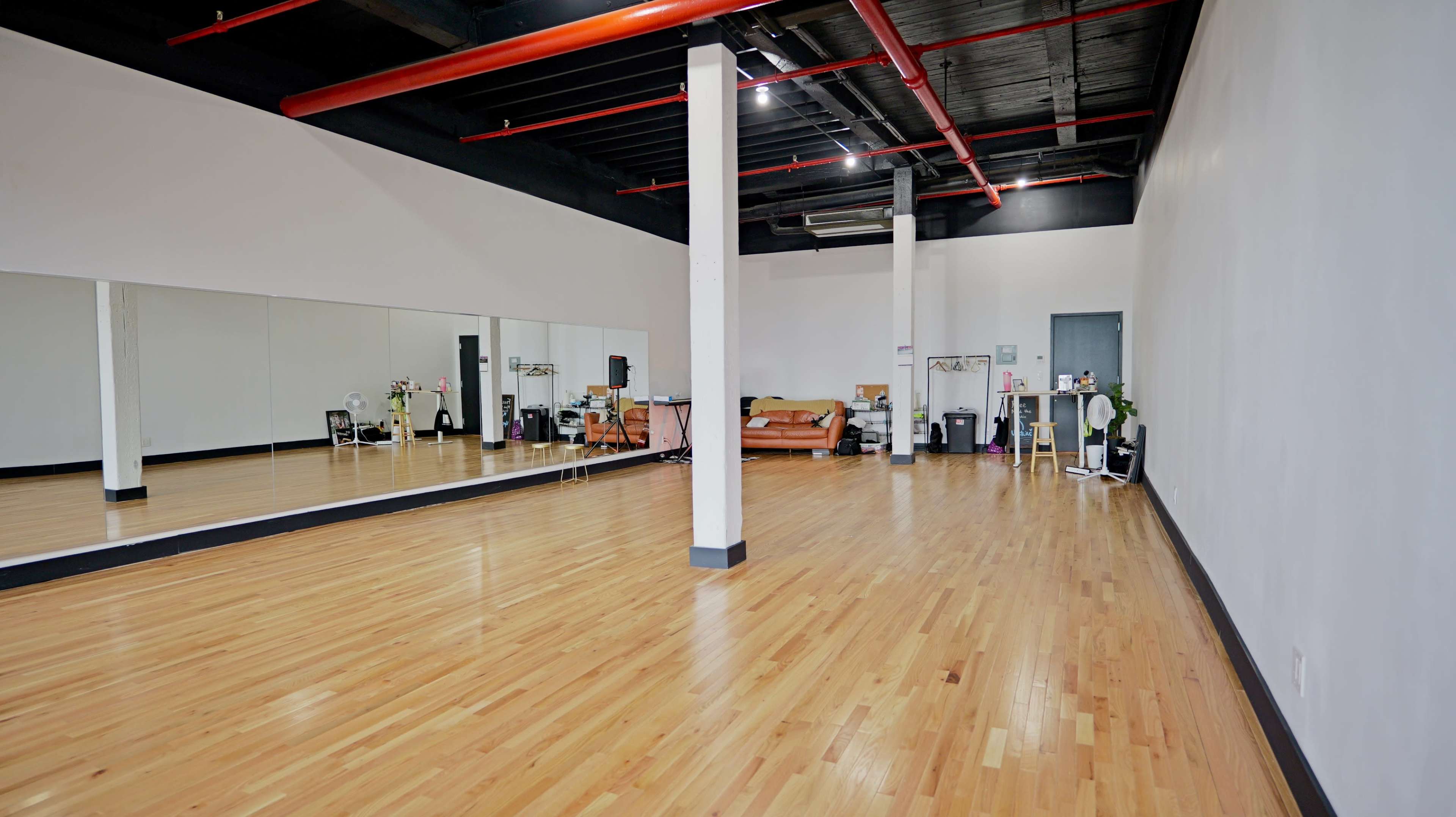 The image shows a spacious studio with a mirrored wall, wooden flooring, and minimal furnishings including a sofa and several plants.