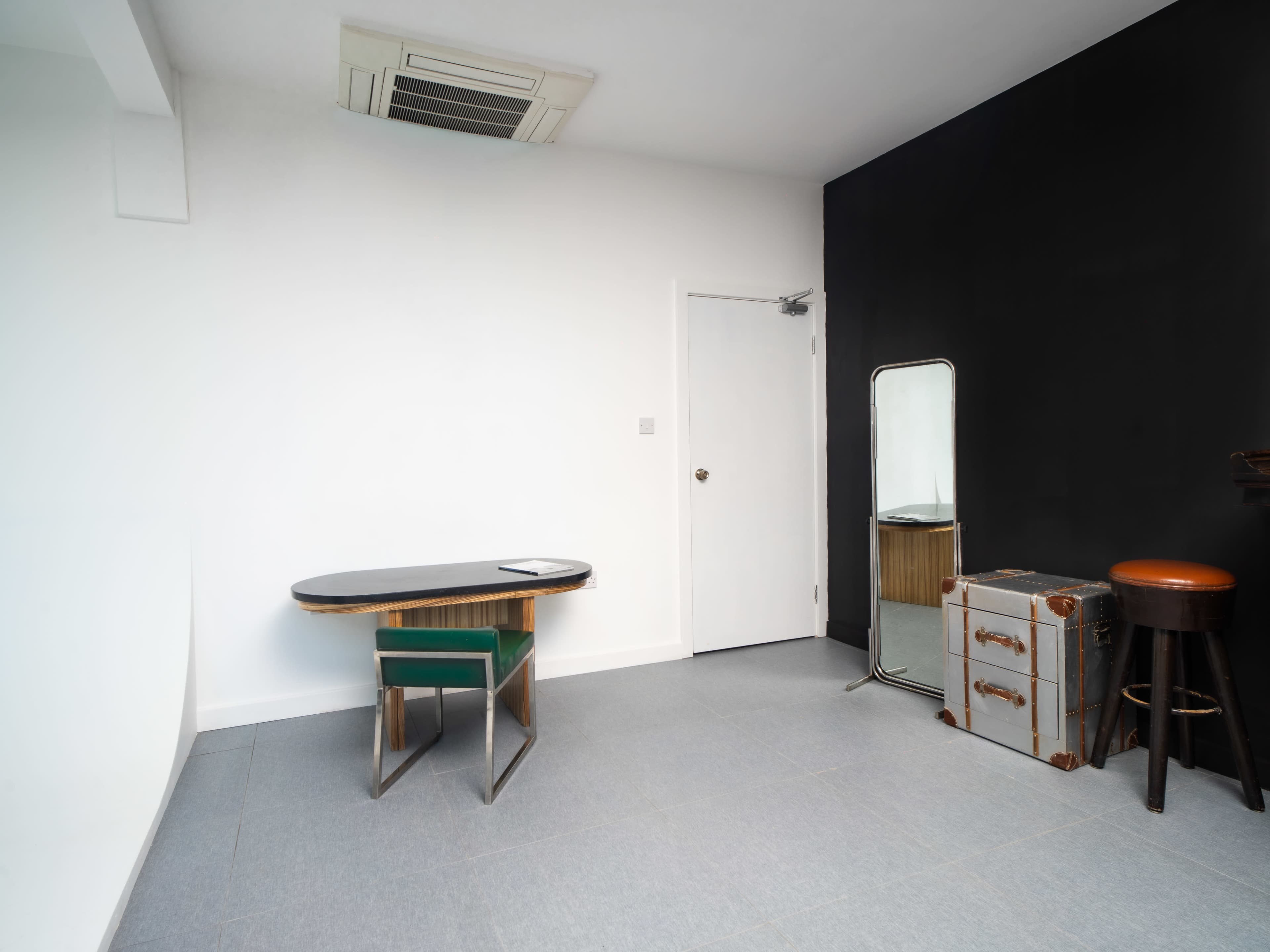 The image shows a minimalist room featuring a desk with a green chair, a mirror, a stool, and a vintage suitcase against a black and white wall.