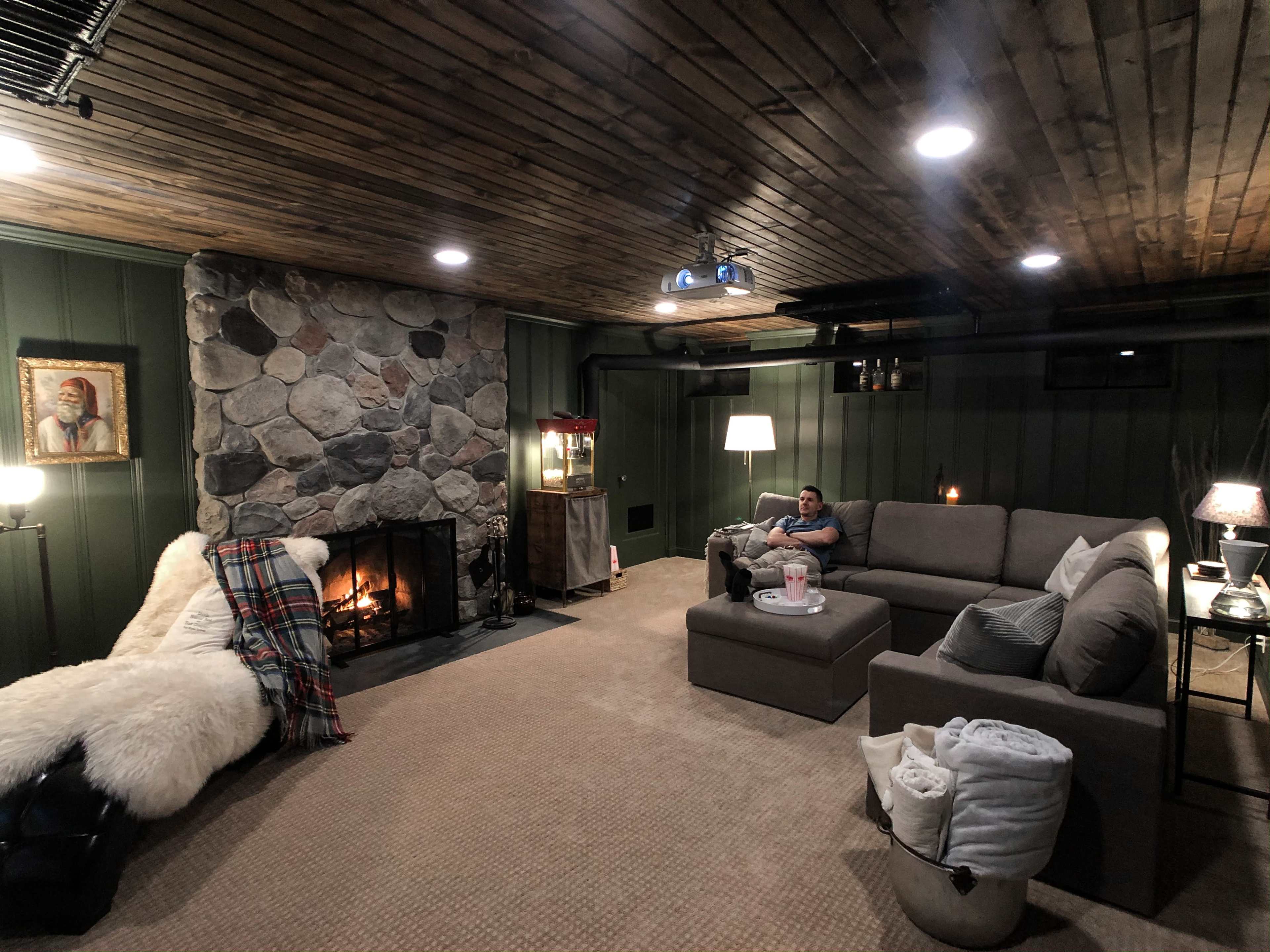 A cozy living room features a stone fireplace, a large sectional sofa, and a projector screen on a wooden ceiling.
