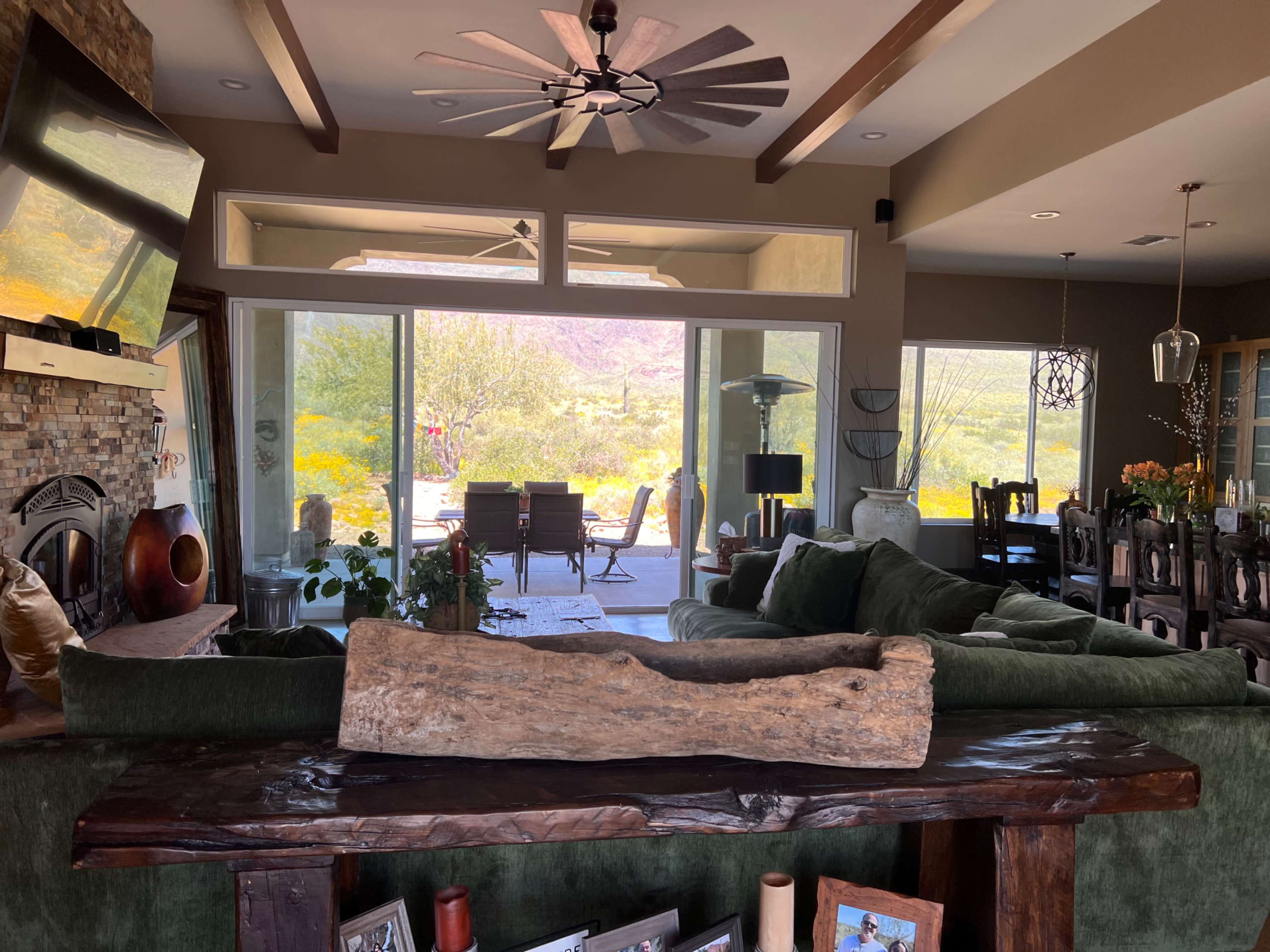 A living room with a green sectional sofa and wooden decor, featuring large windows that open to a view of a patio and outdoor landscape.