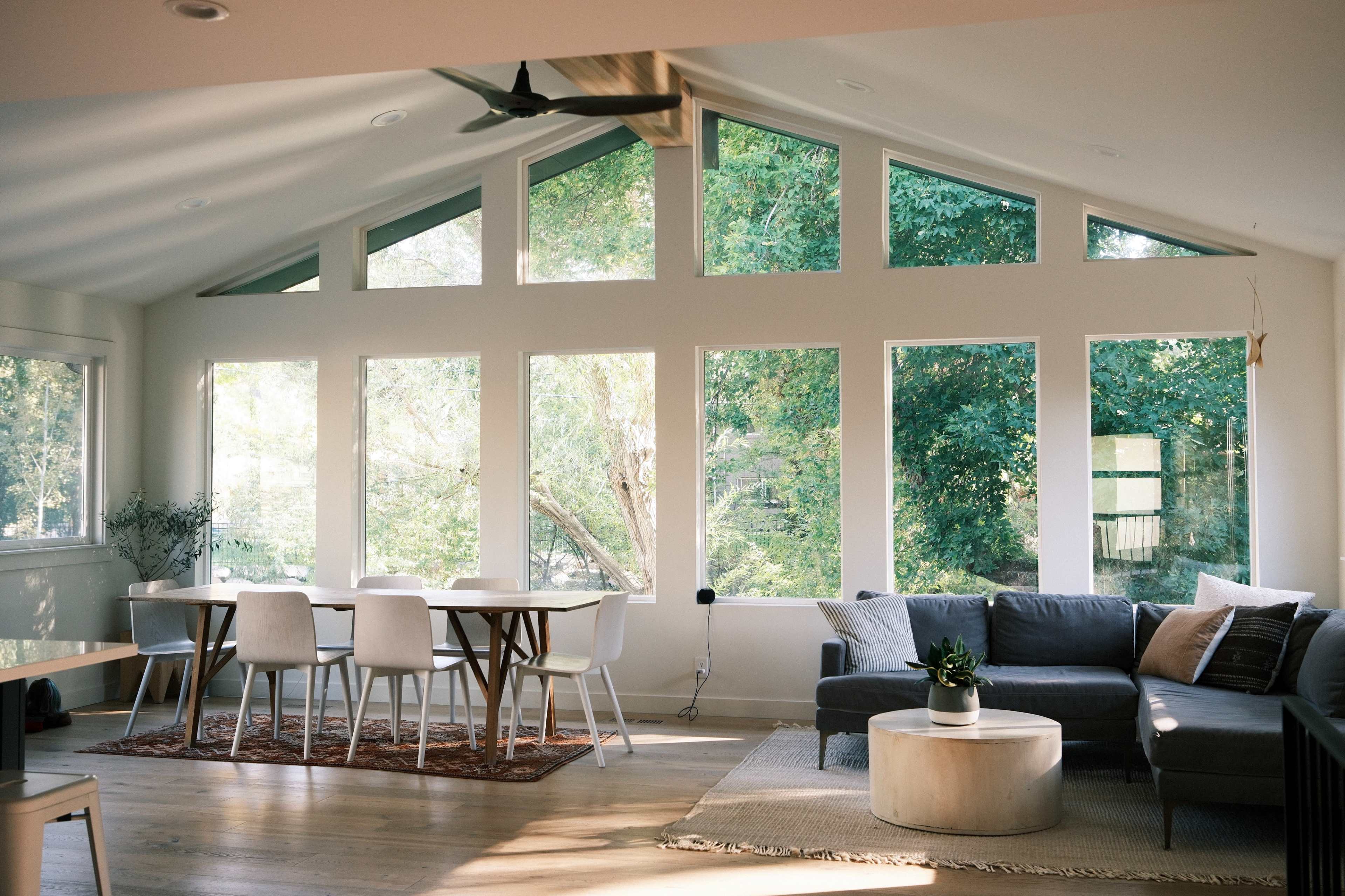 A modern living space features large windows overlooking greenery, a gray sofa, a round coffee table, and a dining area with white chairs.