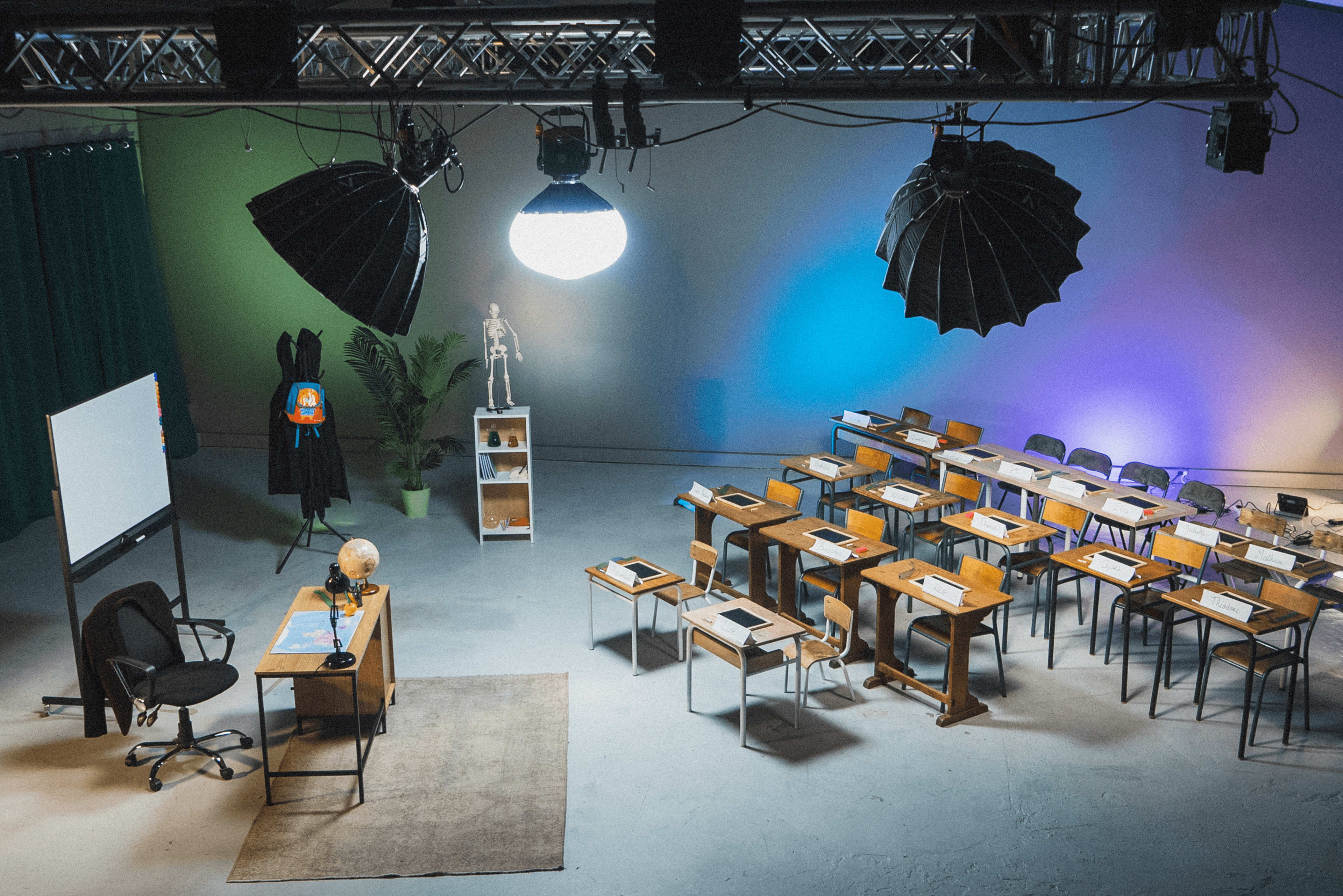 A classroom setting features rows of wooden desks and chairs, a blackboard, a globe, and studio lighting hanging from the ceiling.