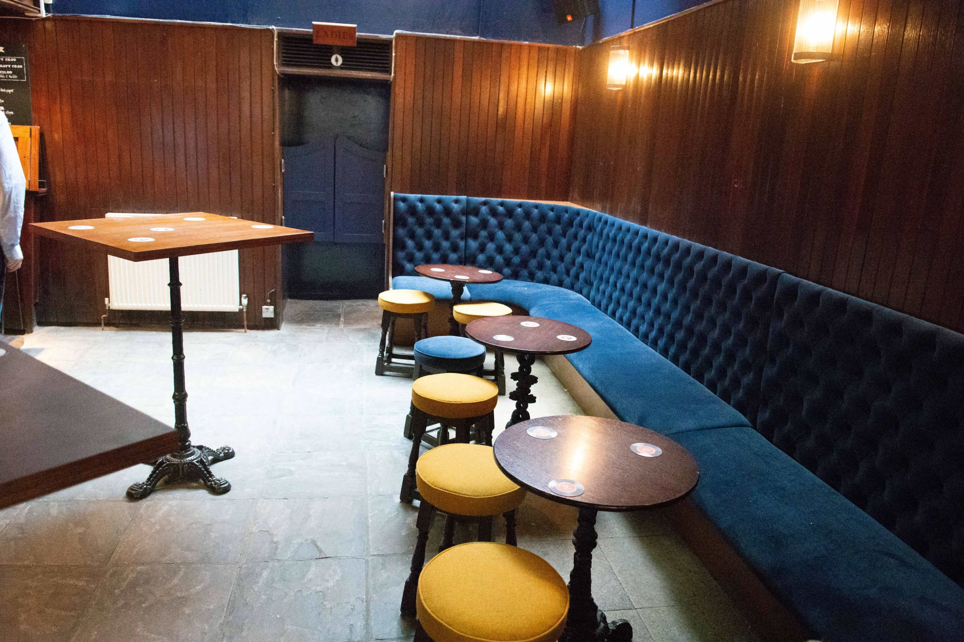 The image shows a cozy bar interior featuring a blue upholstered wall, wooden tables, and stools arranged around a central standing table.