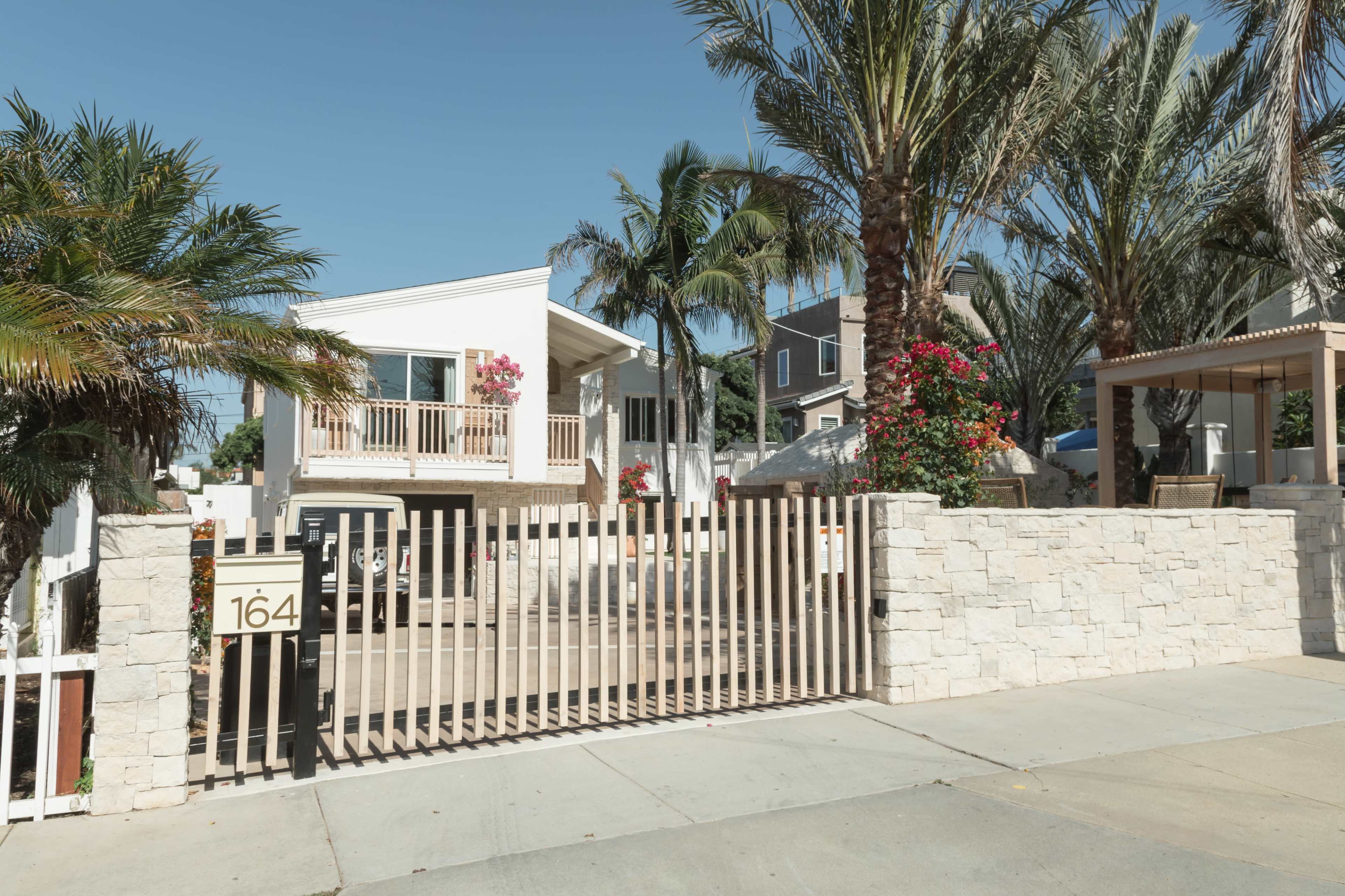 A modern house with a balcony and flowering plants is enclosed by a stone wall and a wooden gate.