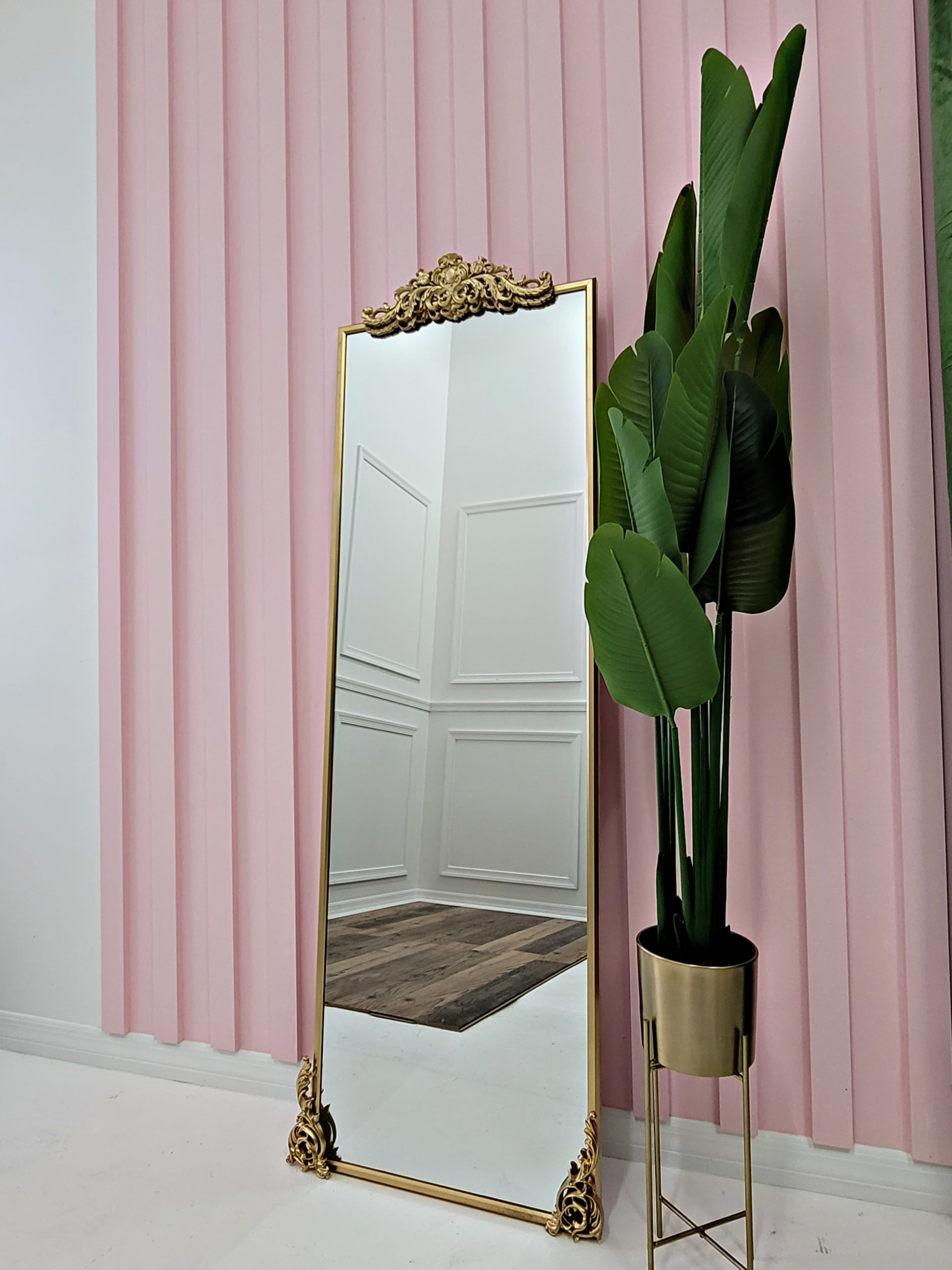A large, ornate mirror stands against a pink wall, reflecting a white room with wainscoting and a wooden floor, beside a potted plant in a gold planter.