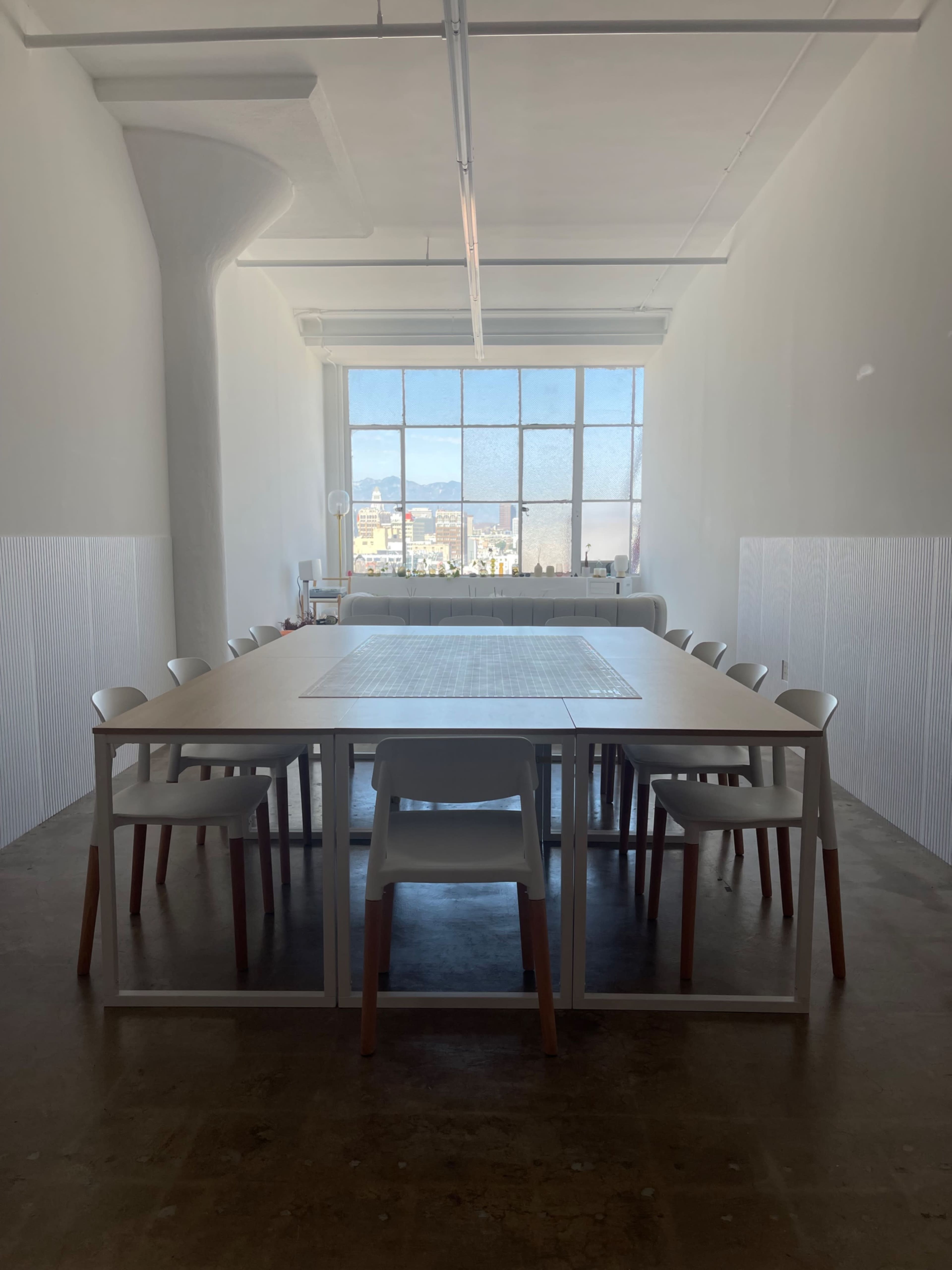 A large, minimalist conference room features a long wooden table surrounded by white chairs, with a window showcasing an urban skyline.