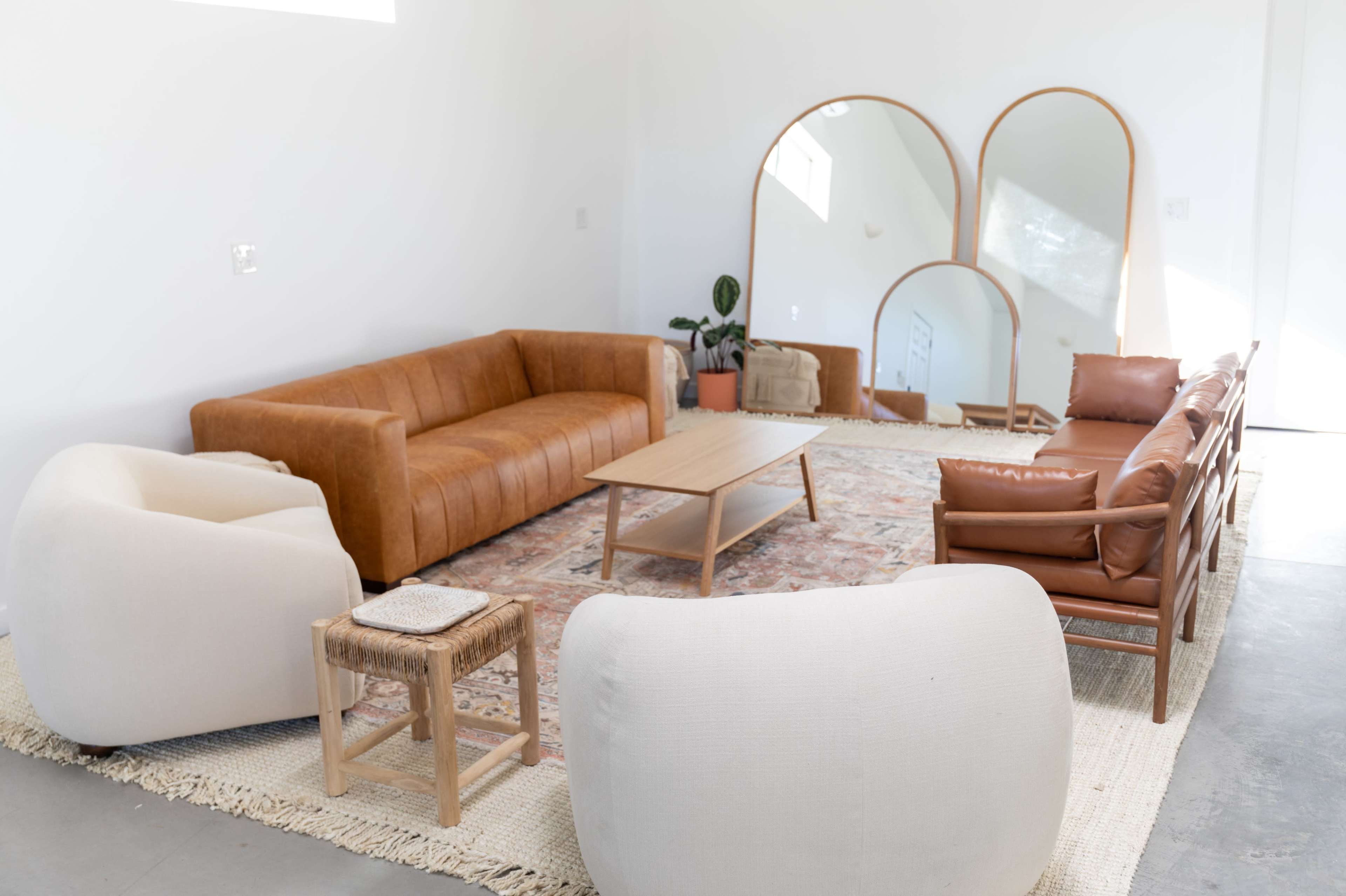 A modern living room features a brown leather sofa, light-colored armchairs, a wooden coffee table, and a patterned area rug, all arranged around large mirrors on a white wall.