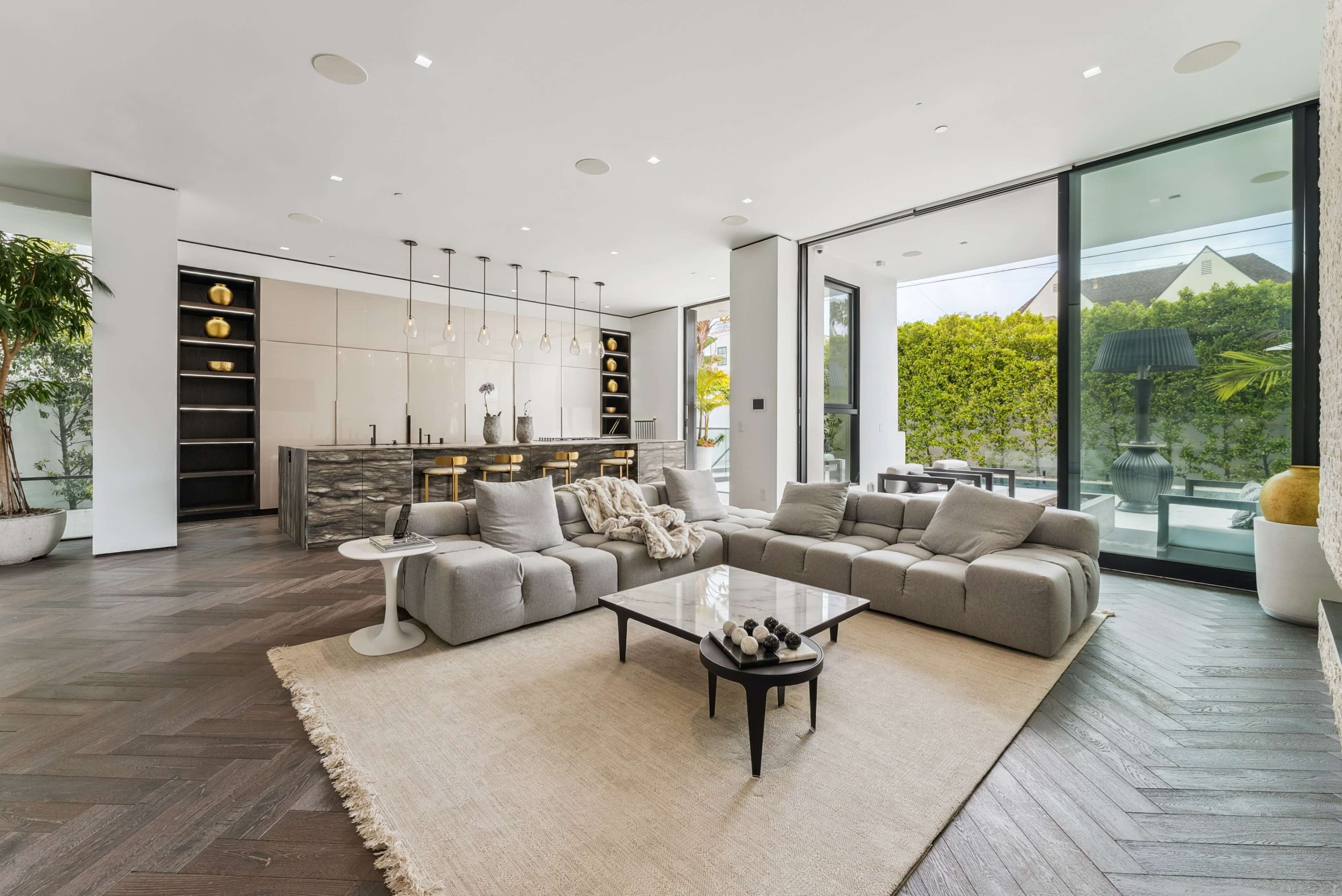 The image shows a modern living room with a large sectional sofa, a coffee table, and an open kitchen area, featuring large windows that provide natural light and a view of outdoor greenery.