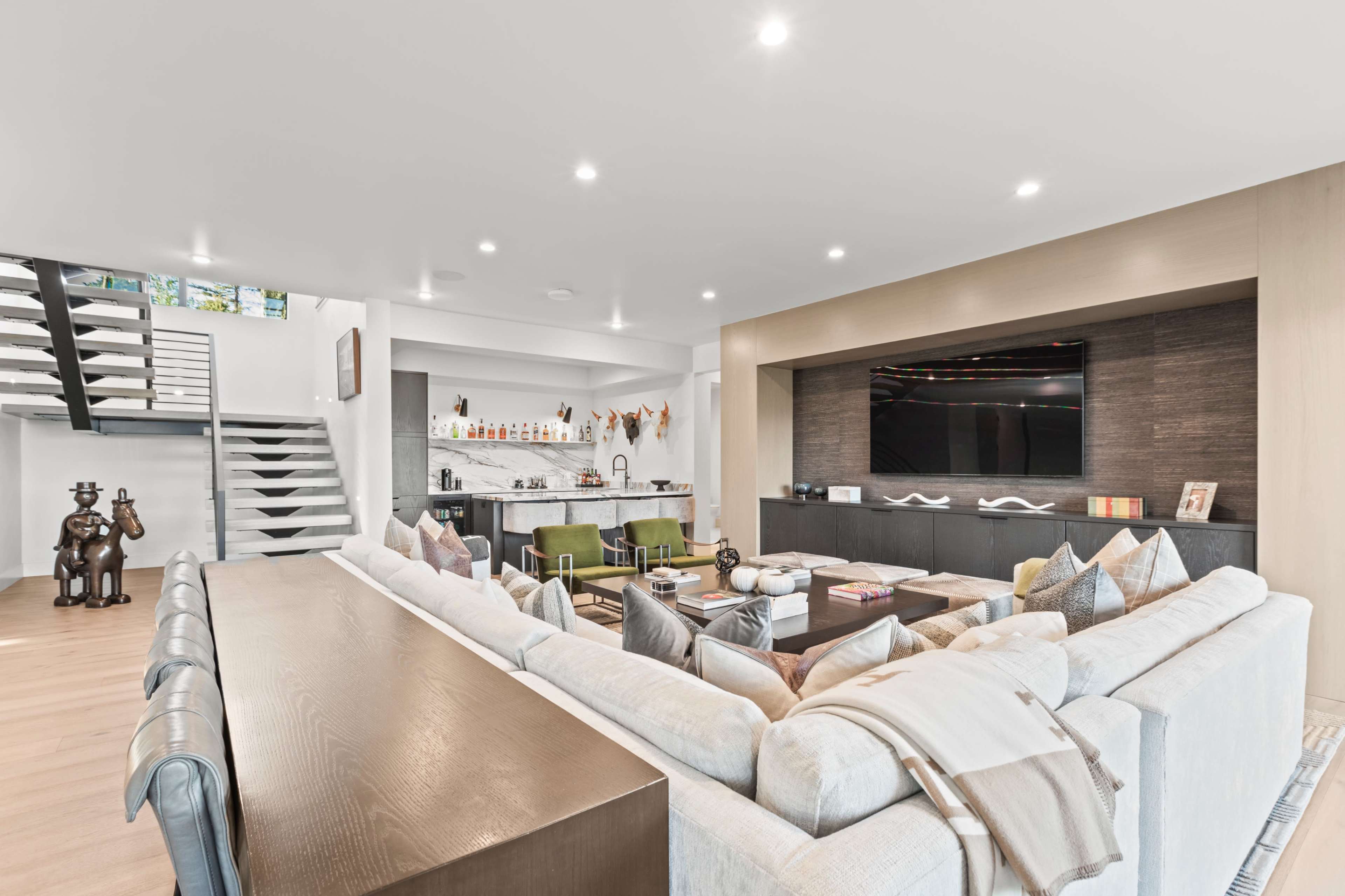 A modern living room features a large sectional sofa, a coffee table, and a wall-mounted television, with a staircase leading to an upper level.