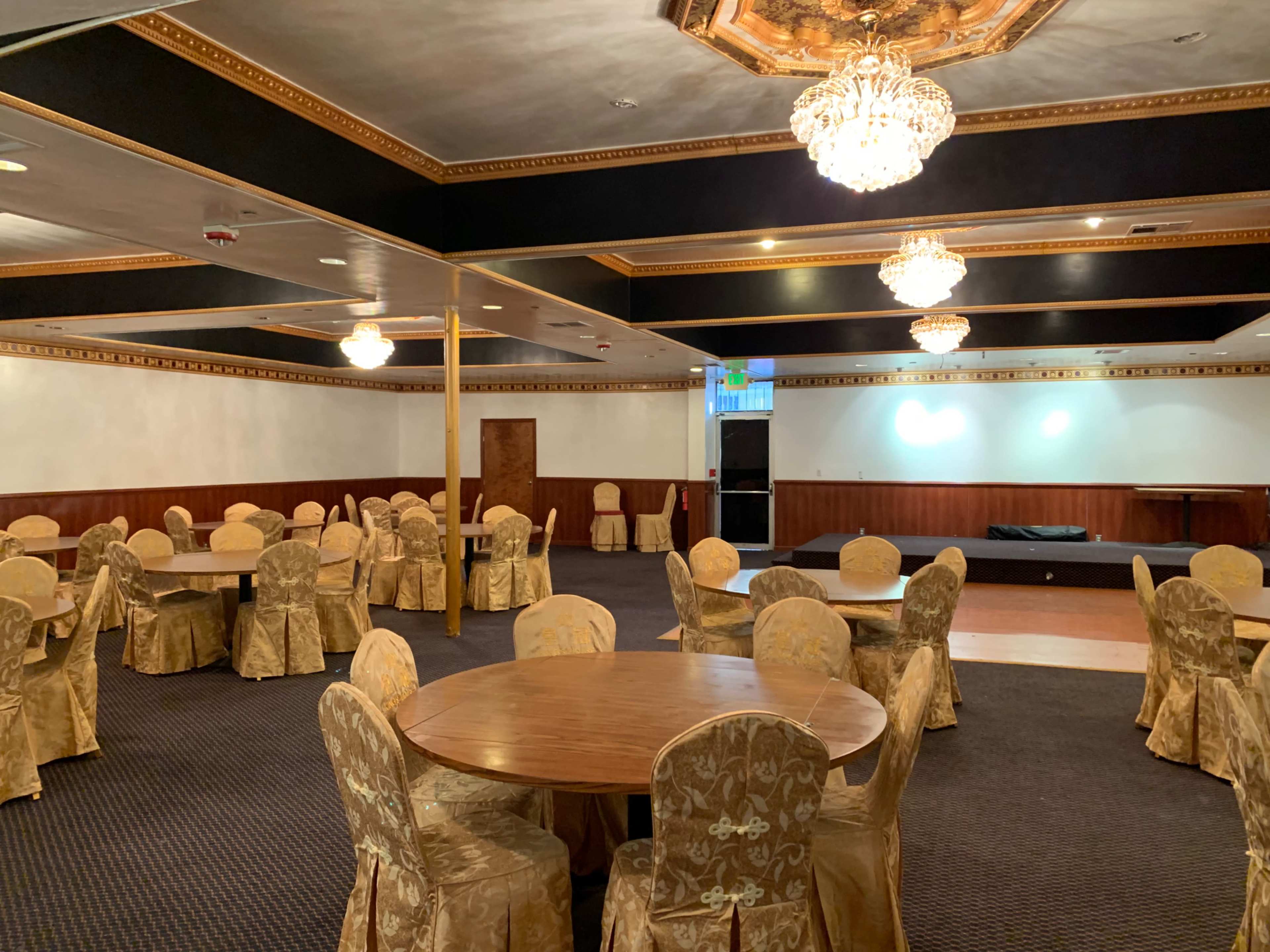 A large banquet hall furnished with round tables and gold-covered chairs, illuminated by several chandeliers.