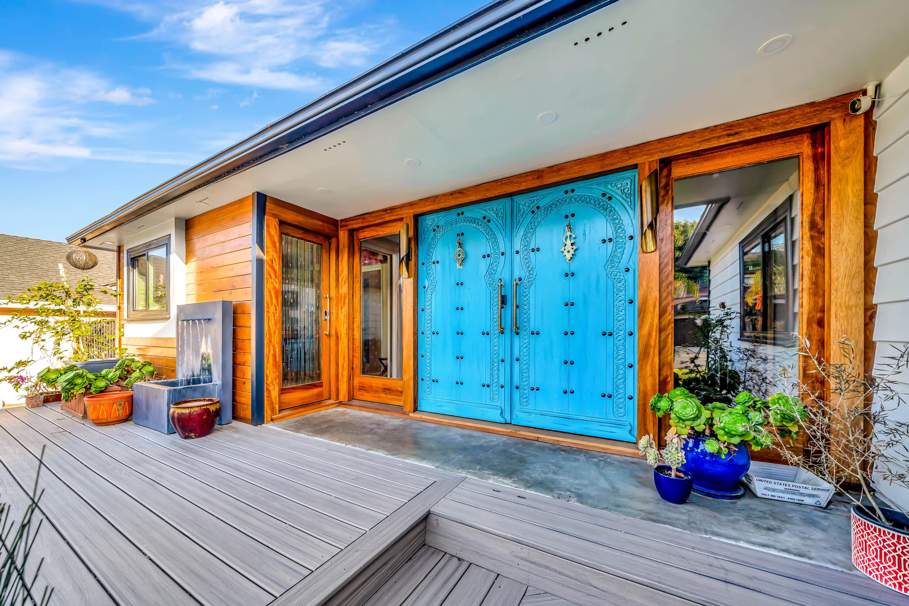 A house entrance featuring large blue doors with intricate designs, surrounded by wooden accents and potted plants on a wooden deck.