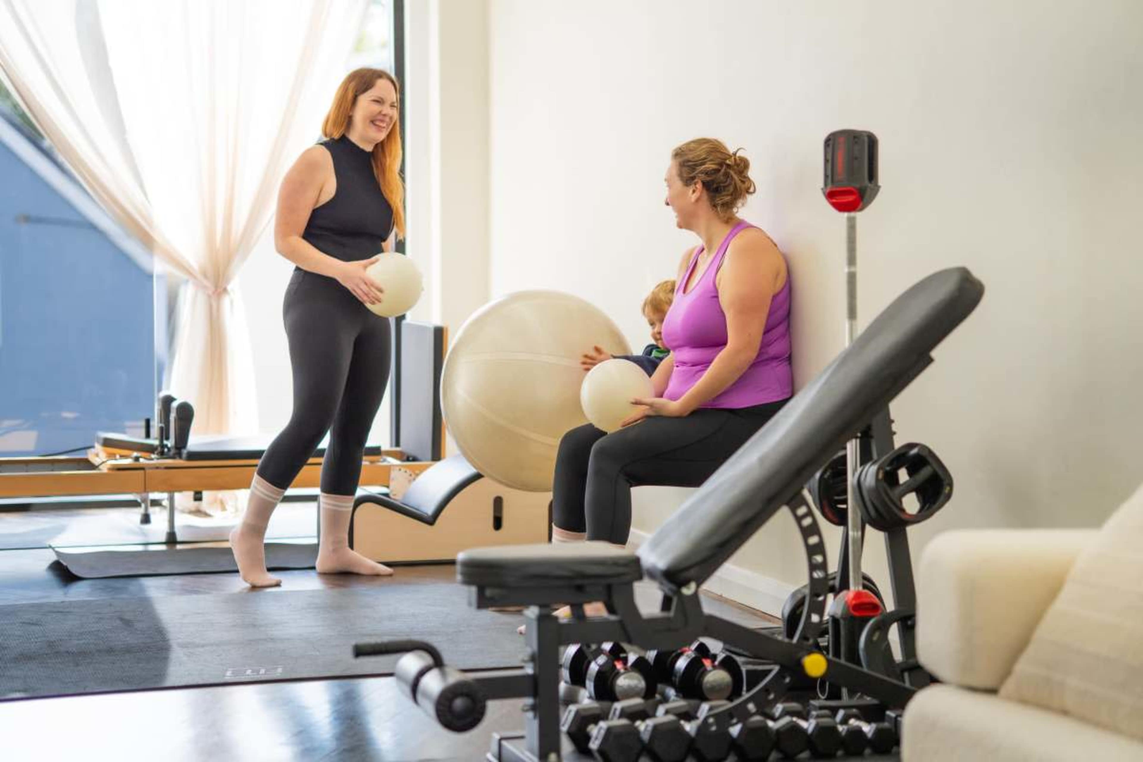 Two women are engaged in a conversation while holding exercise balls in a fitness studio equipped with workout equipment.