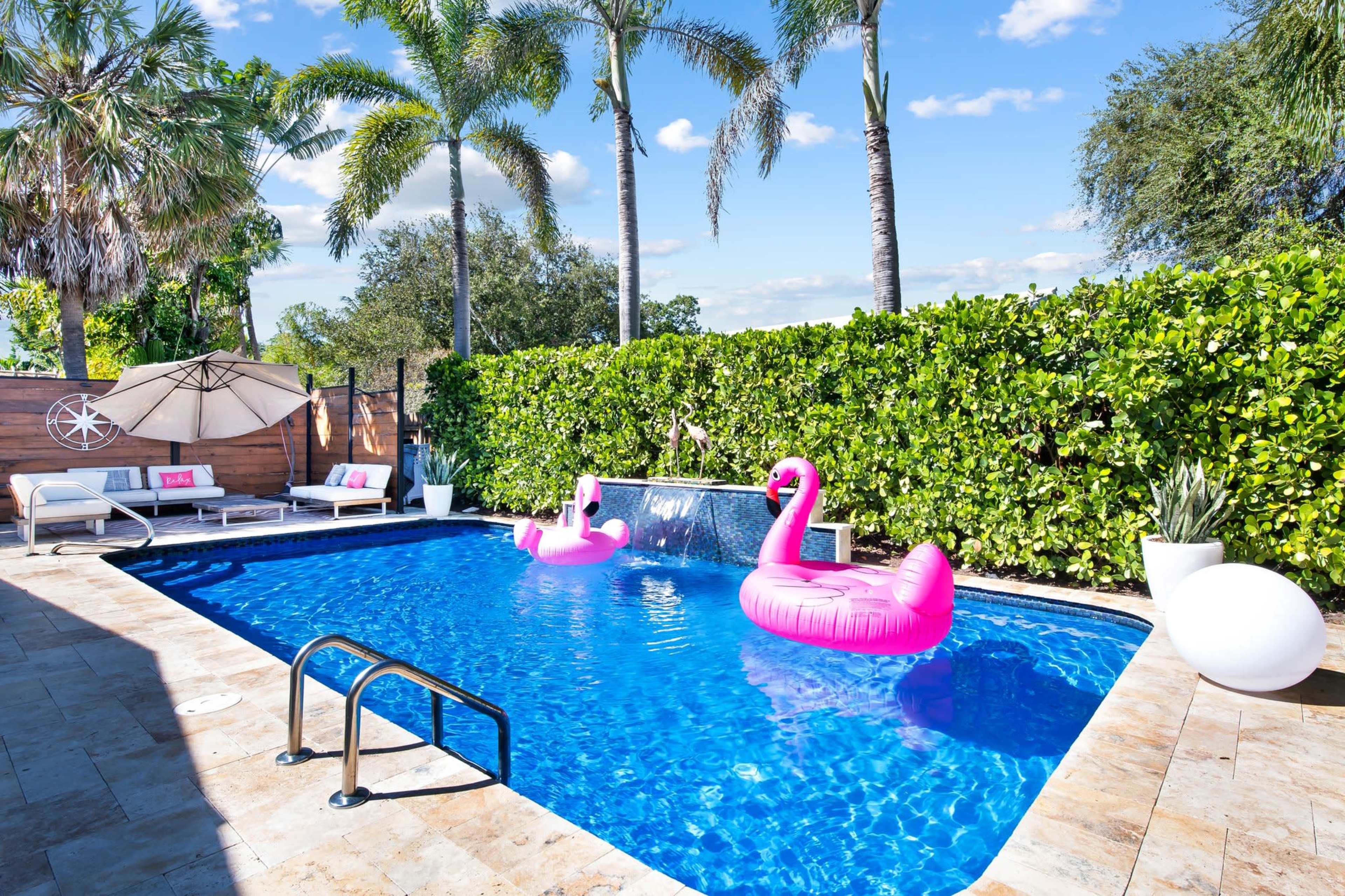 A backyard swimming pool surrounded by green hedges, two pink flamingo floaties, lounge chairs, and a shaded area with an umbrella.