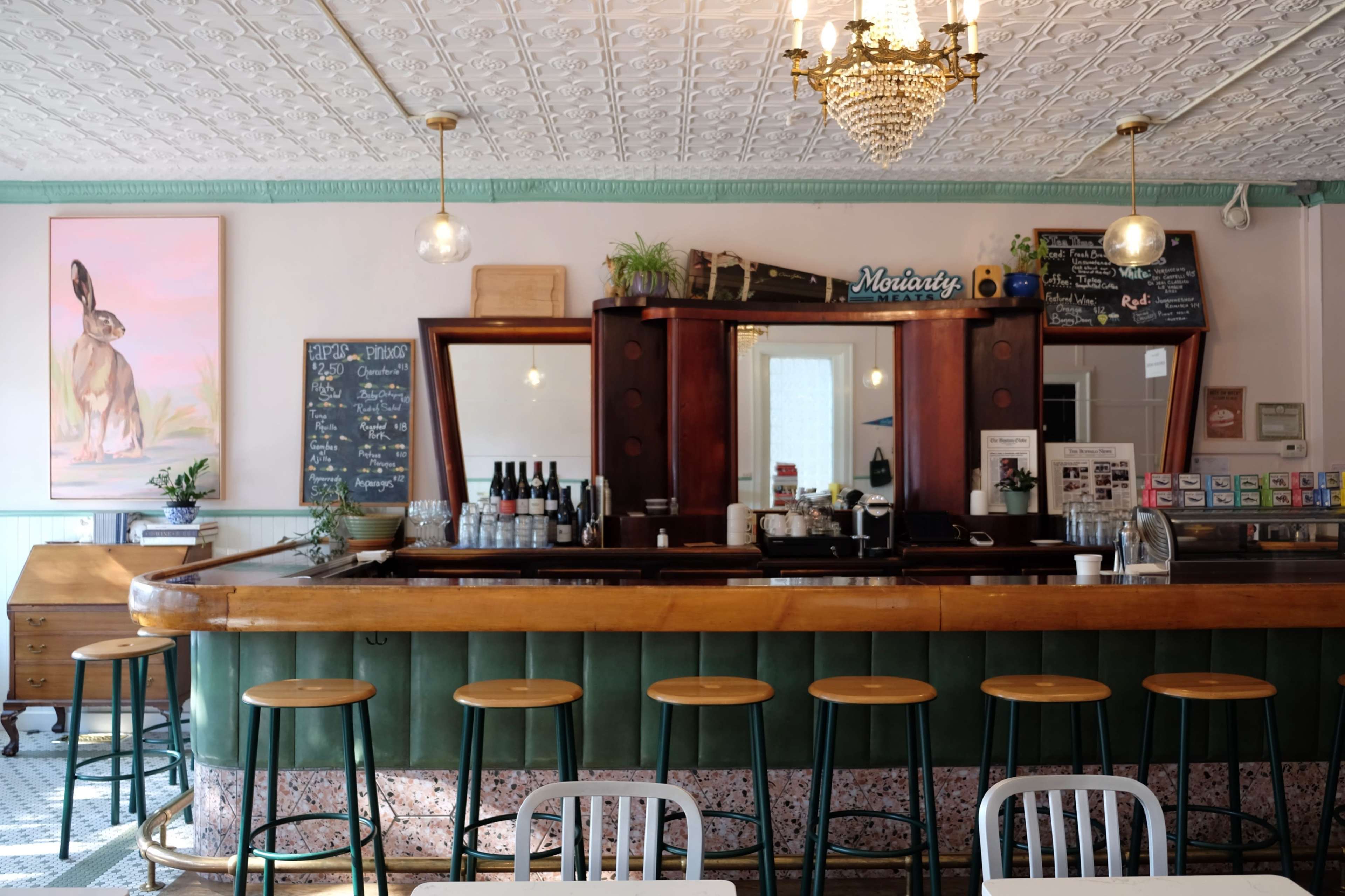 A vintage-style bar with a wooden counter, green leather seating, and a large mirror, adorned by plants and light fixtures.