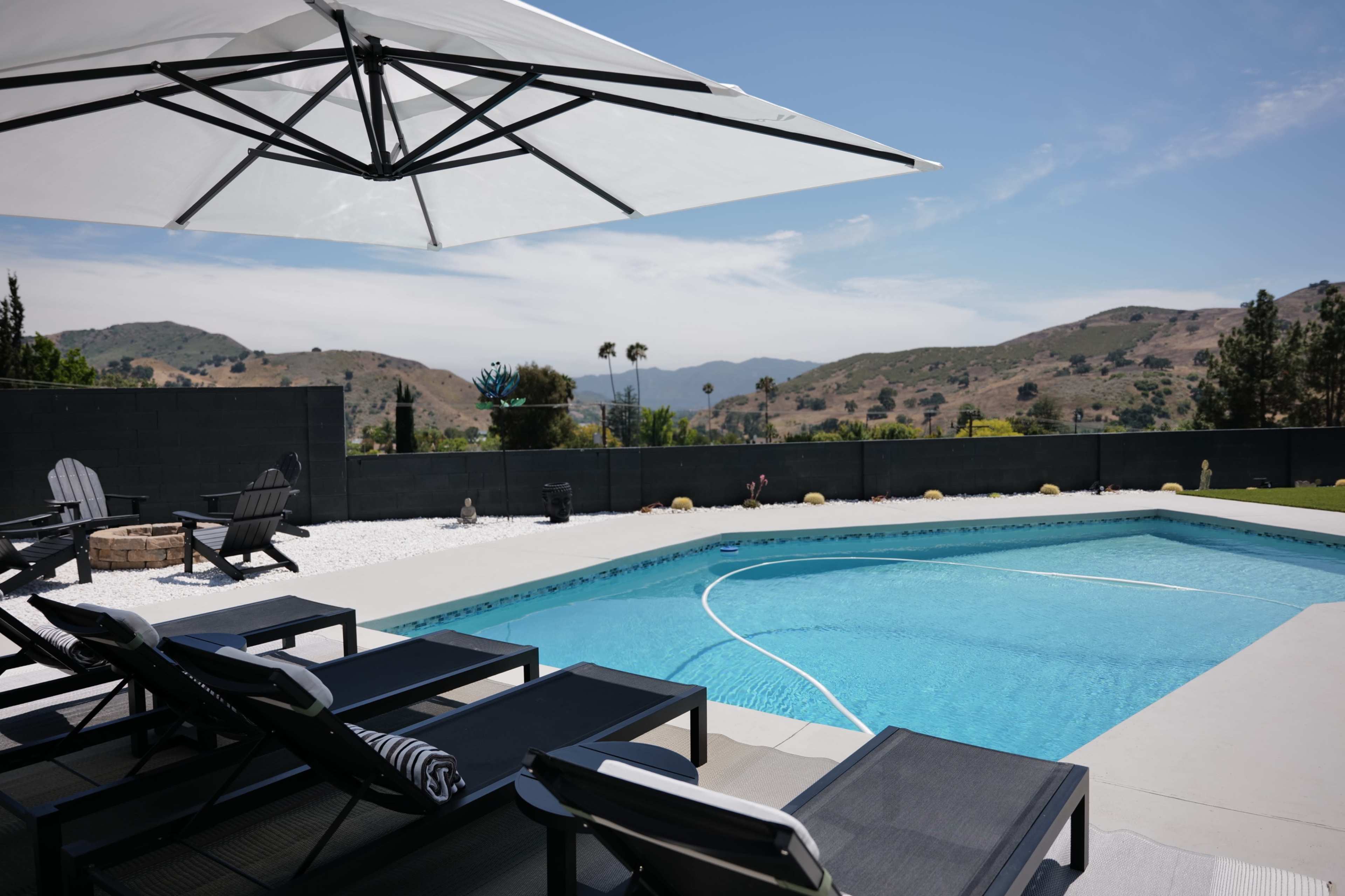 A swimming pool with black lounge chairs under a large umbrella is set against a backdrop of rolling hills and a clear sky.