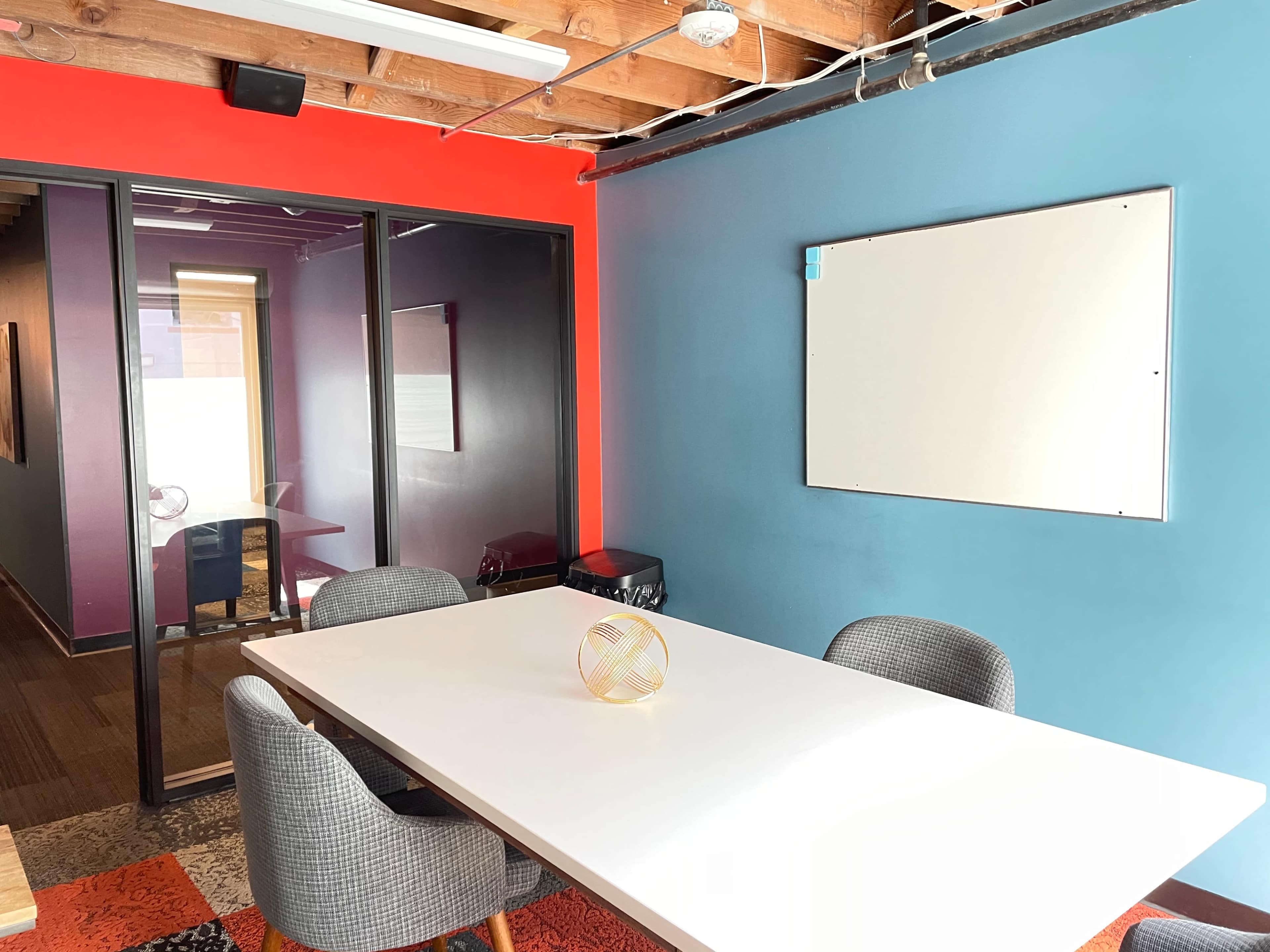 A meeting room with a white table, gray chairs, and a colorful wall, featuring a whiteboard and a glass wall partition.