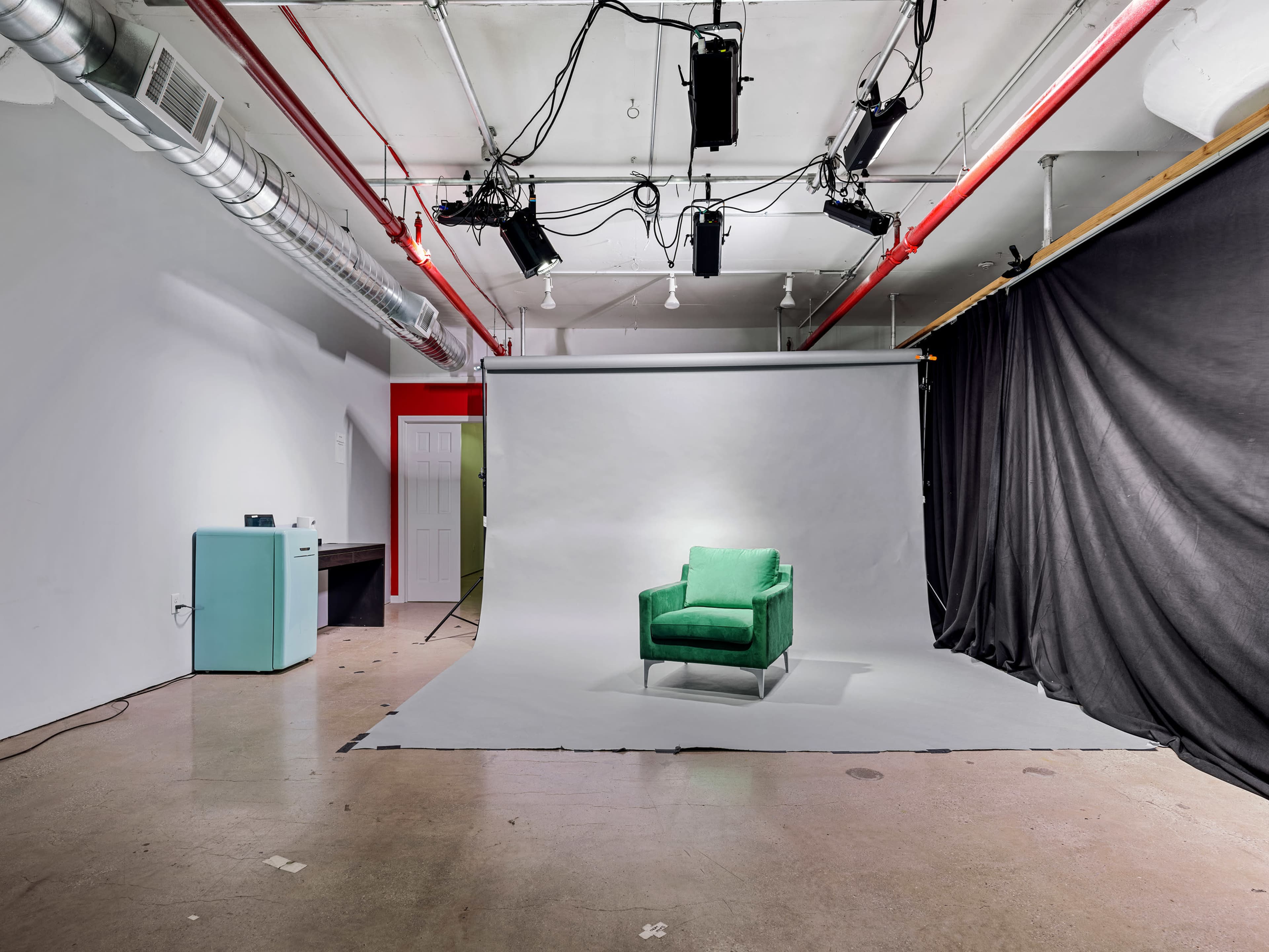 The image shows a photography studio featuring a green armchair positioned on a light backdrop, with overhead lighting and dark curtains in the background.