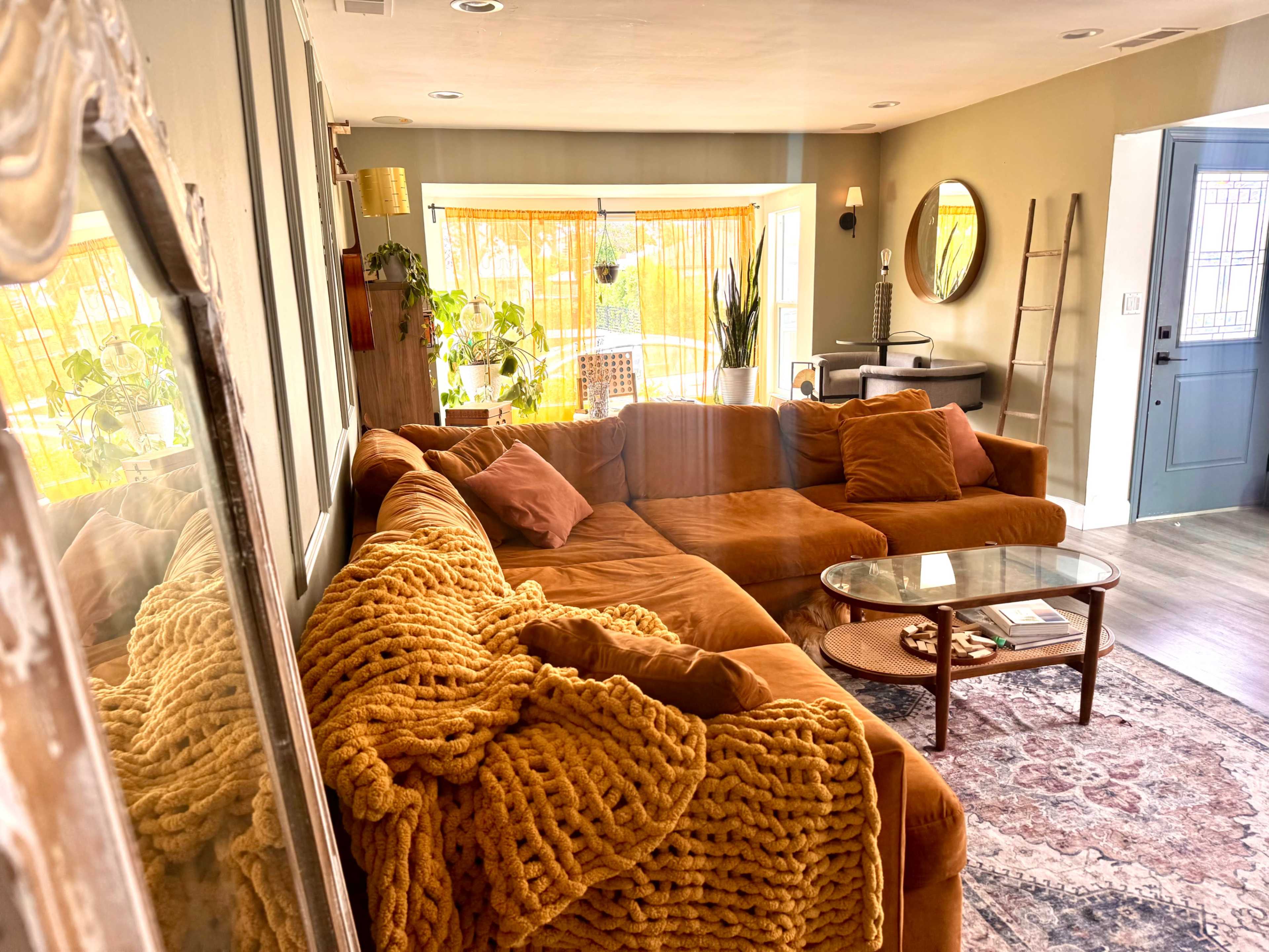 A spacious living room features a large, orange sectional sofa with a textured blanket draped over it, a glass coffee table, and natural light streaming in through large windows.