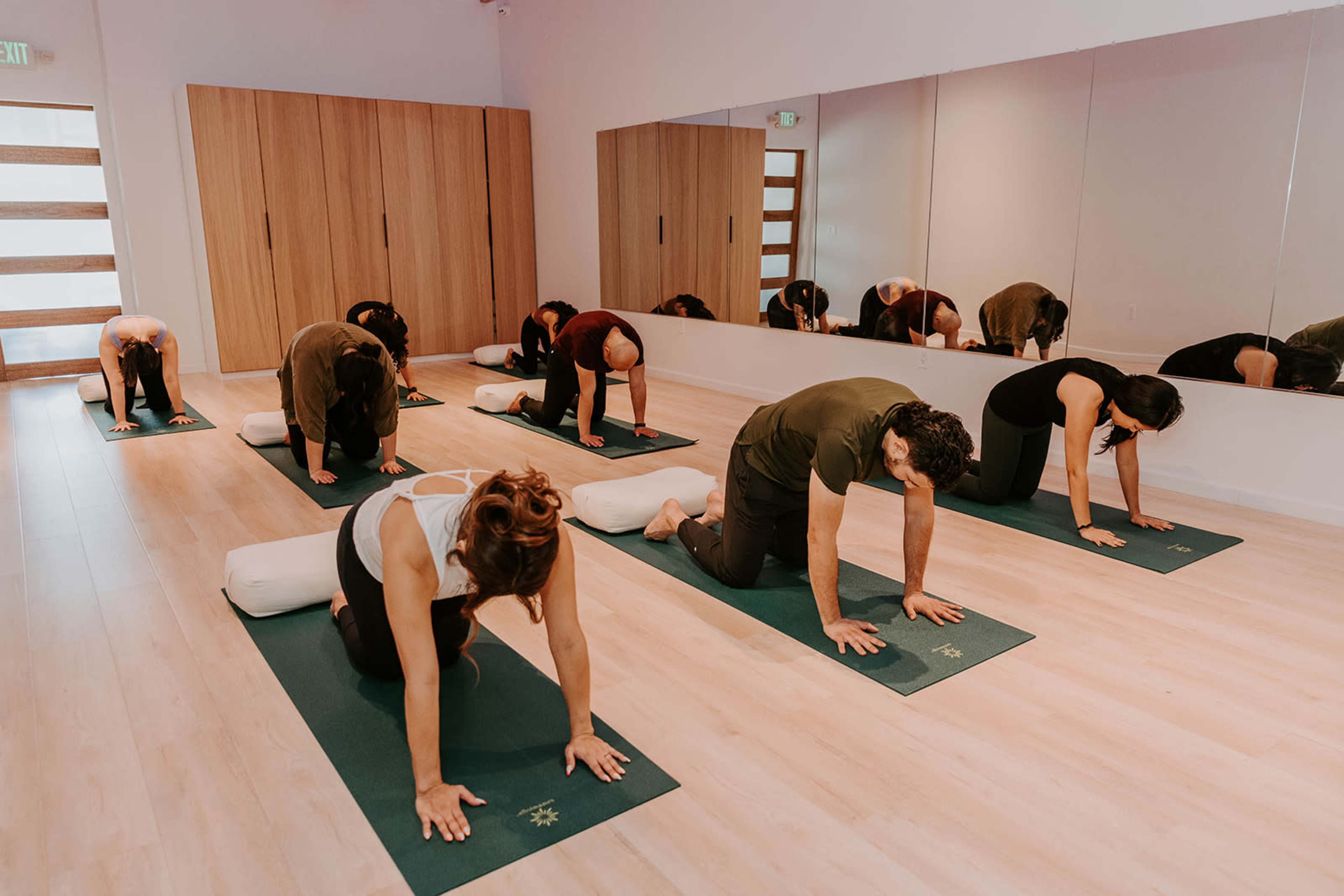 A group of people is practicing yoga on mats in a well-lit studio with mirrors and wooden paneling.