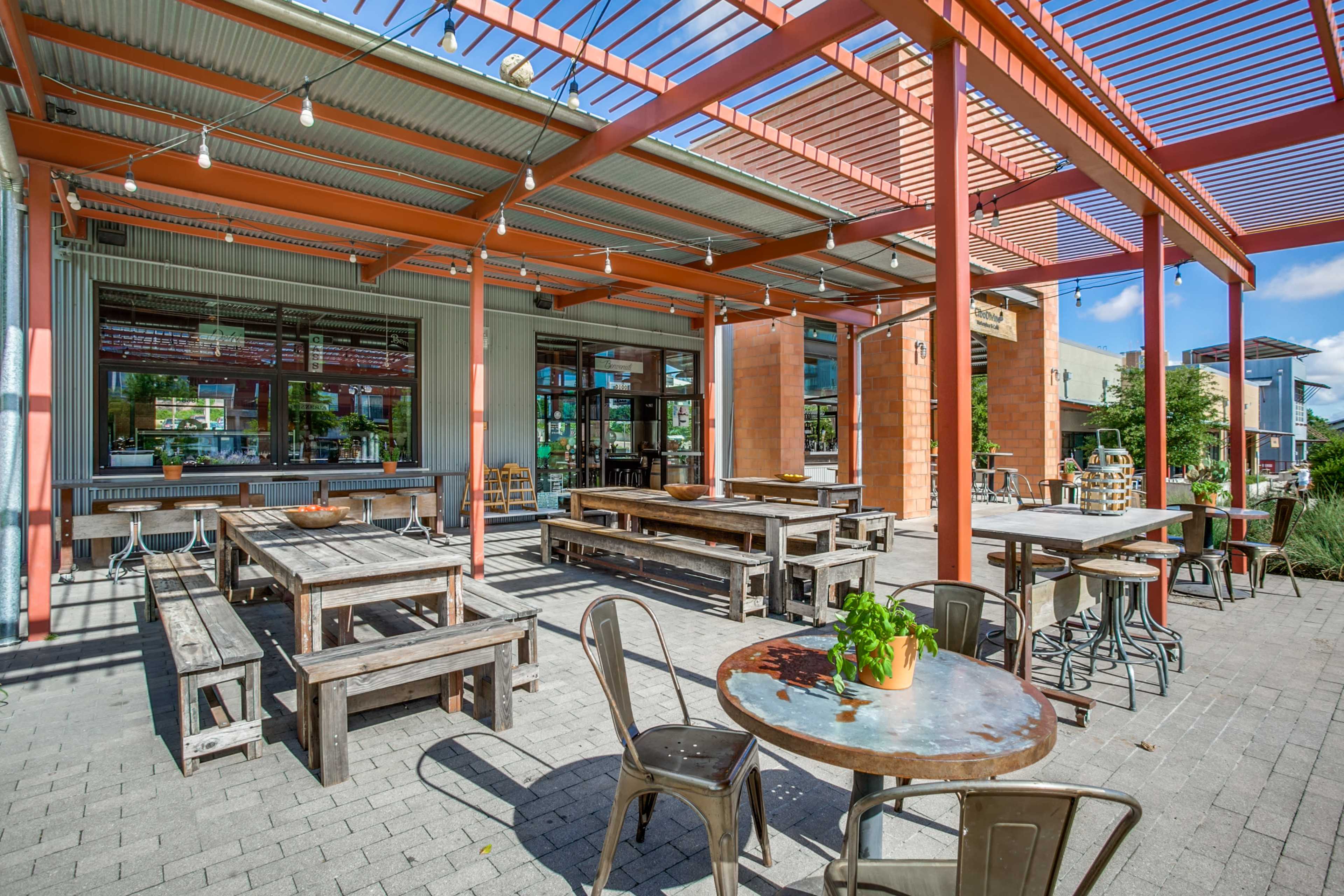 An exterior dining area with wooden tables and metal chairs under a red framed awning.