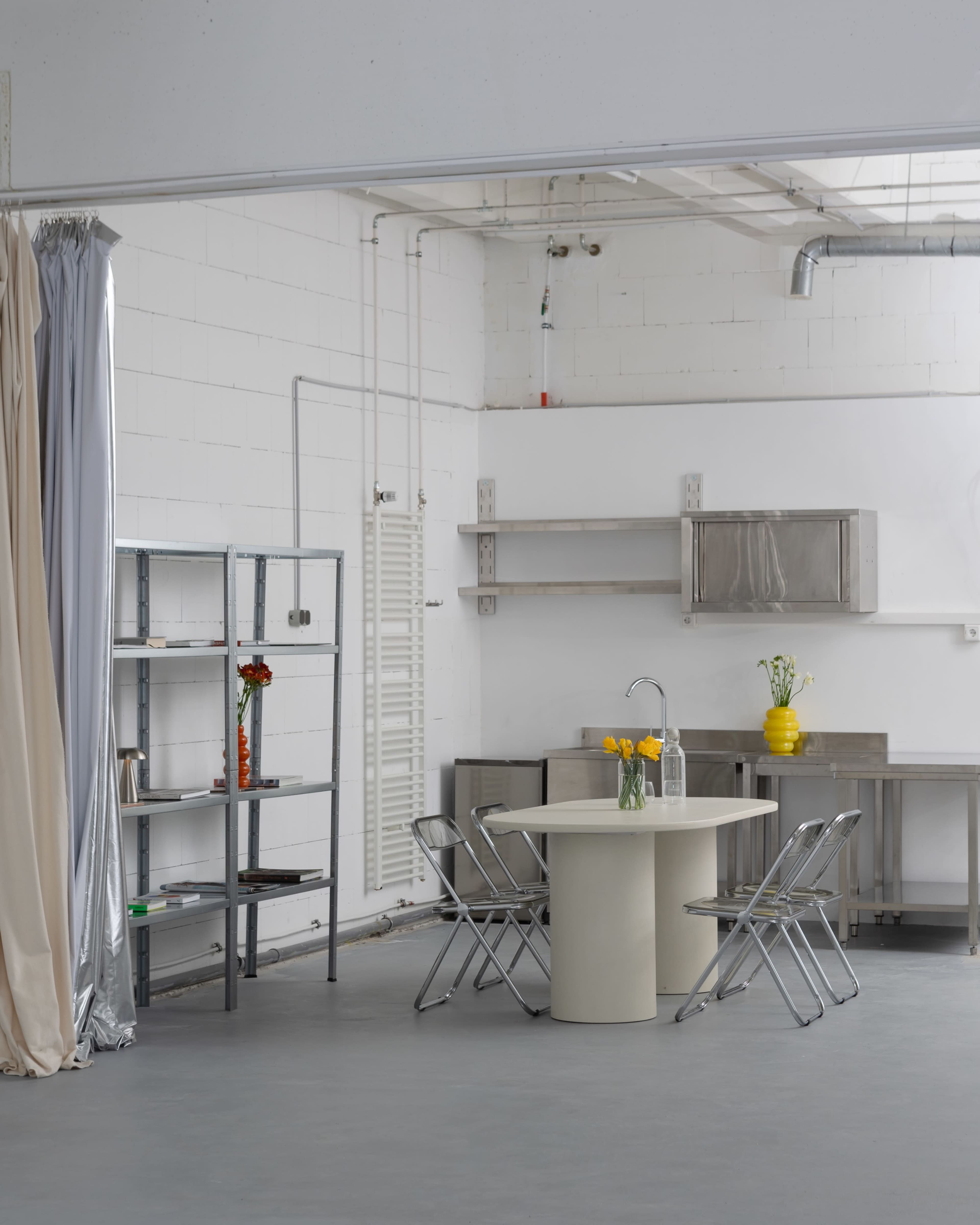 The image shows a minimalist kitchen space featuring a round table with four folding chairs, a stainless steel shelf, and a few decorative items.