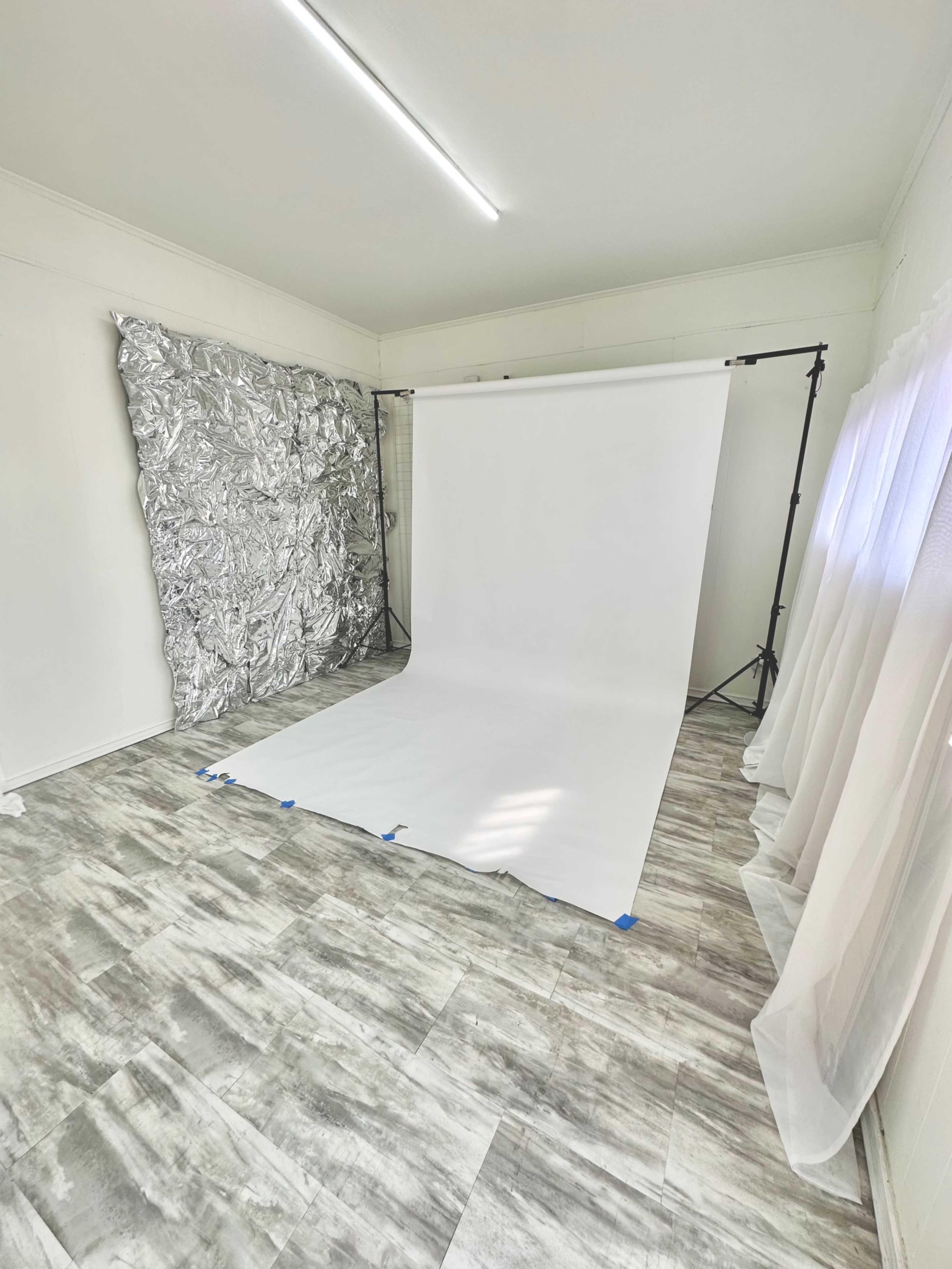 A photography studio setup featuring a white backdrop, a silver reflective surface on the wall, and gray tiled flooring.