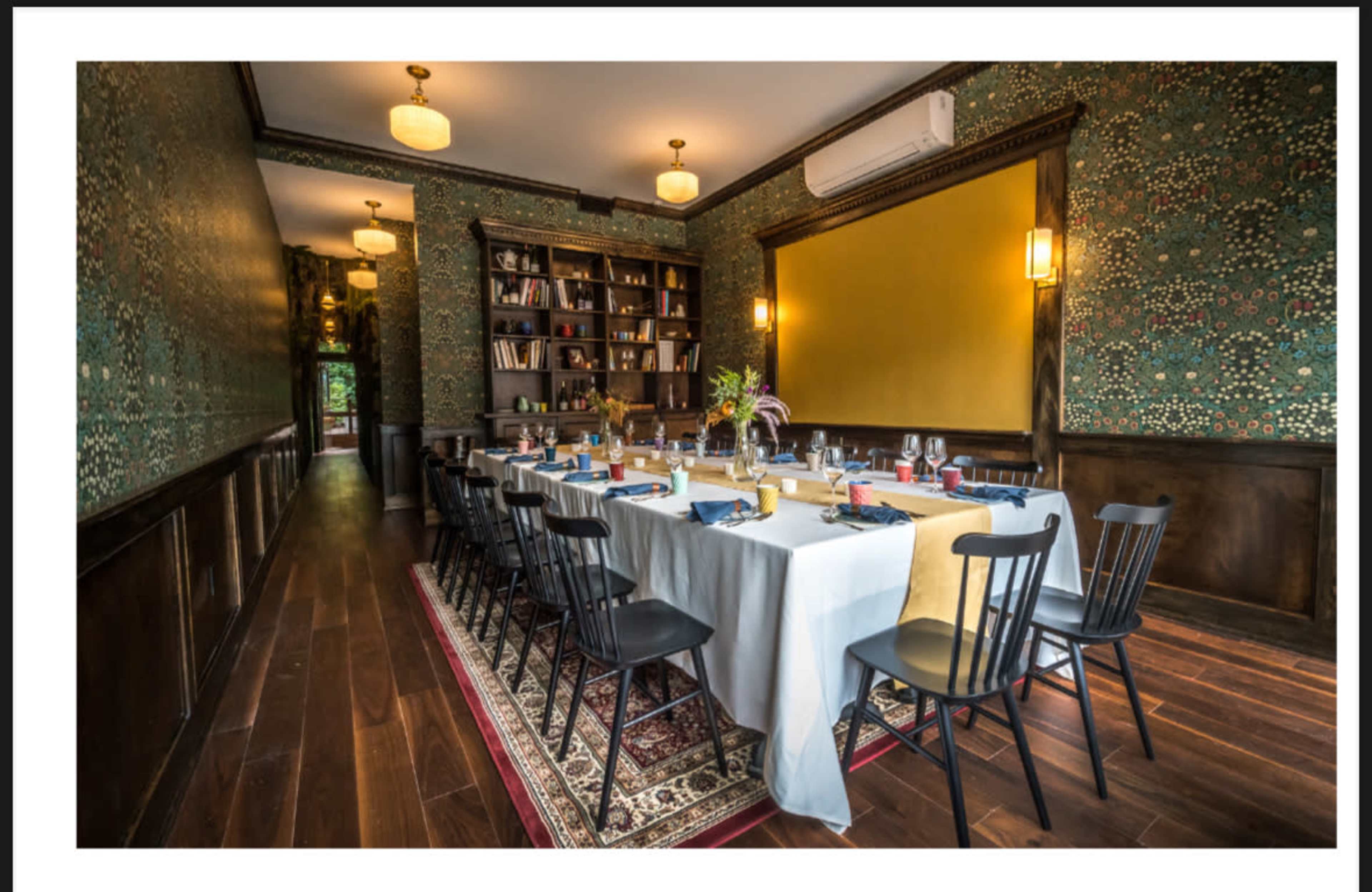 A long dining table is set in a richly decorated room with wallpaper, bookshelves, and warm lighting.