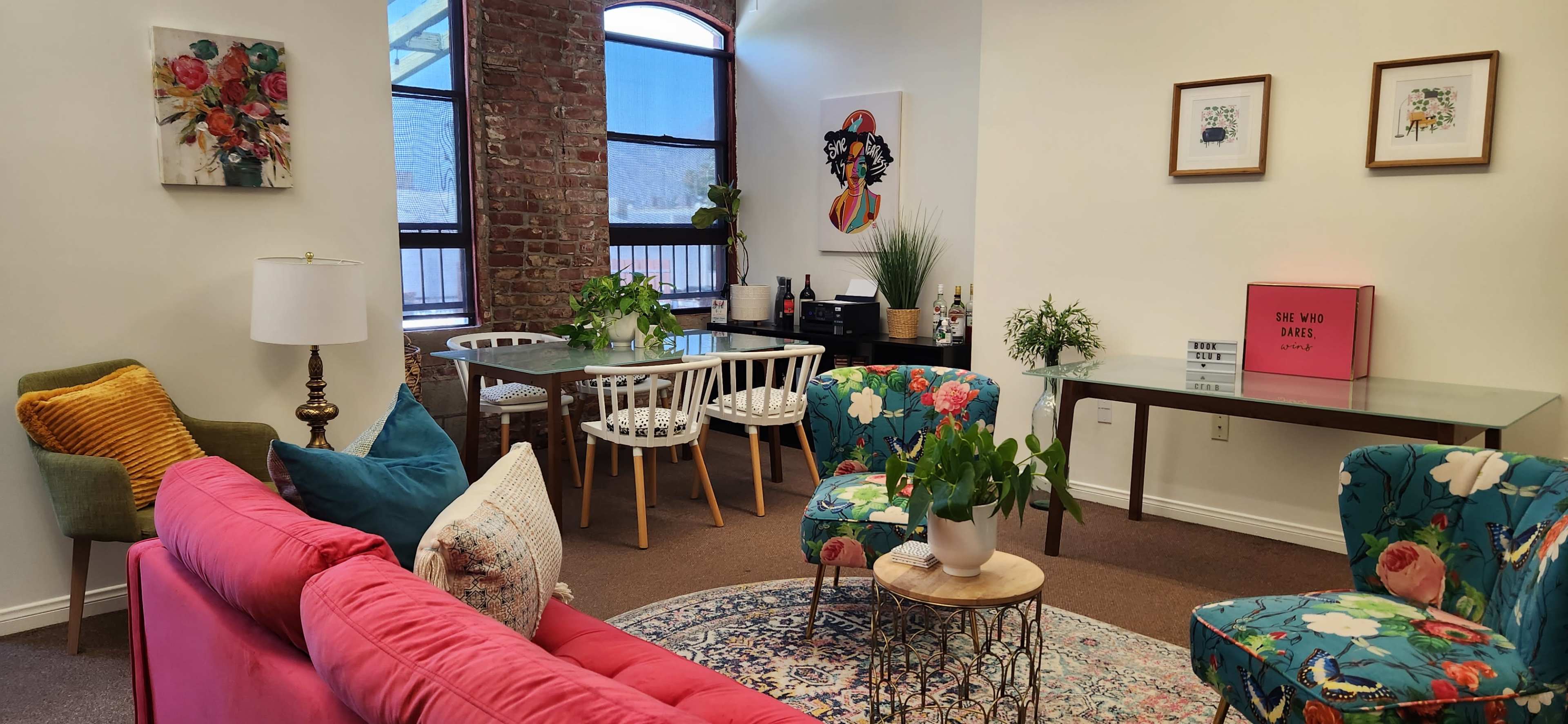 A cozy room with a pink sofa, floral chairs, a dining table, and various plants against a backdrop of exposed brick and large windows.