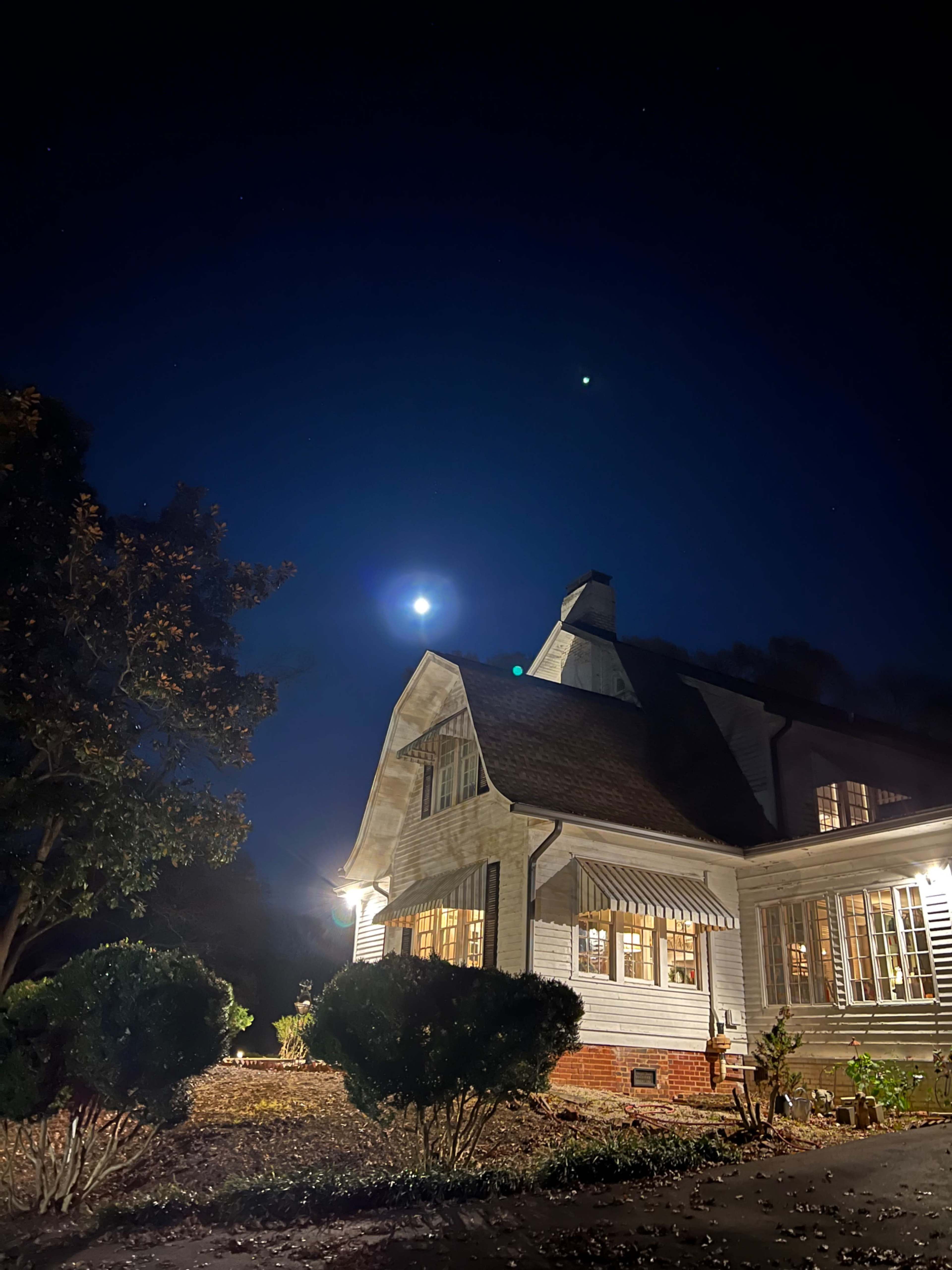 A house with illuminated windows stands under a night sky, featuring a bright moon and a planet visible above.